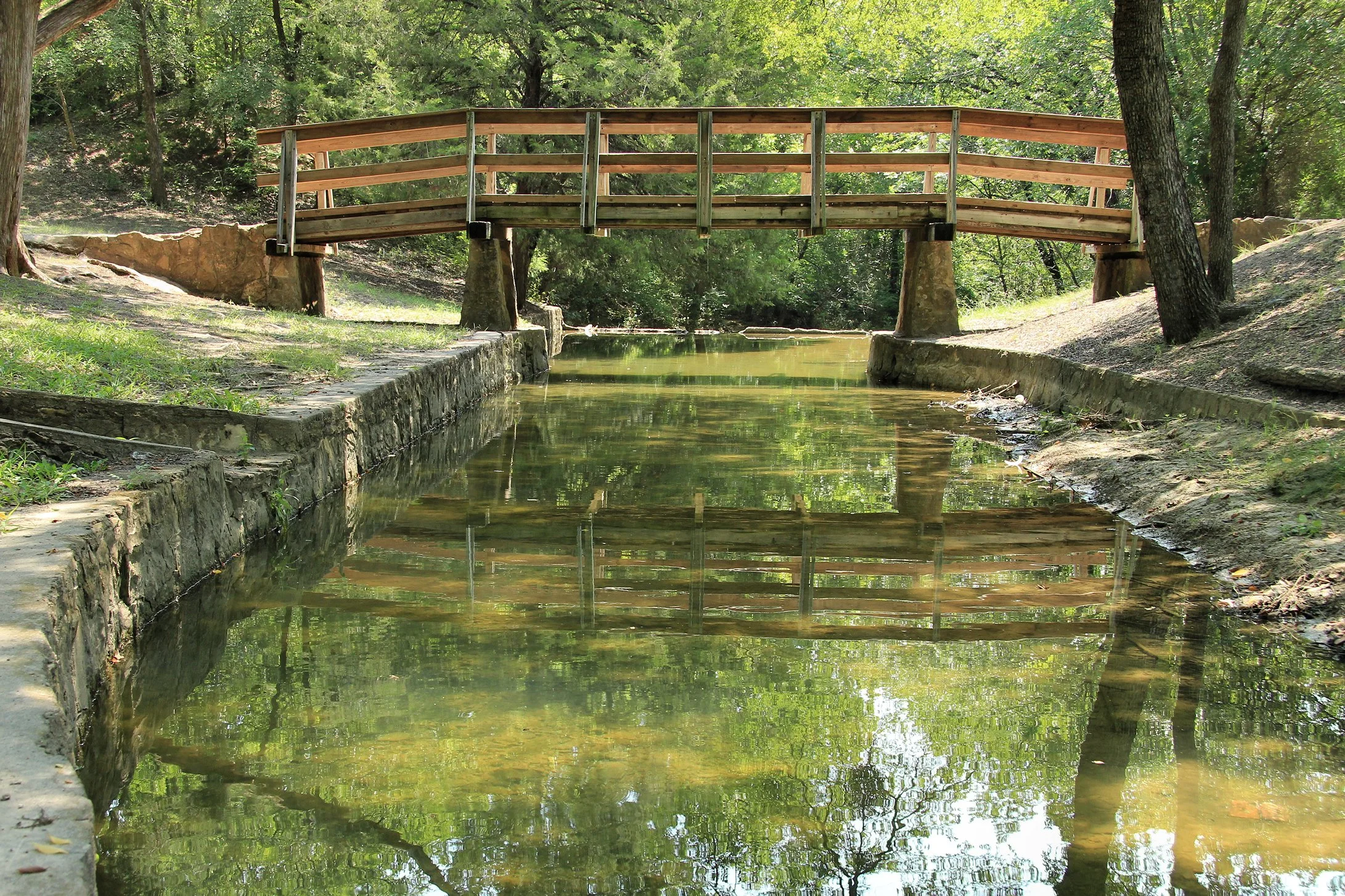 A small wooden bridge crossing over a narrow, calm stream in a wooded park, with green trees and grass on both sides in Coppell Texas