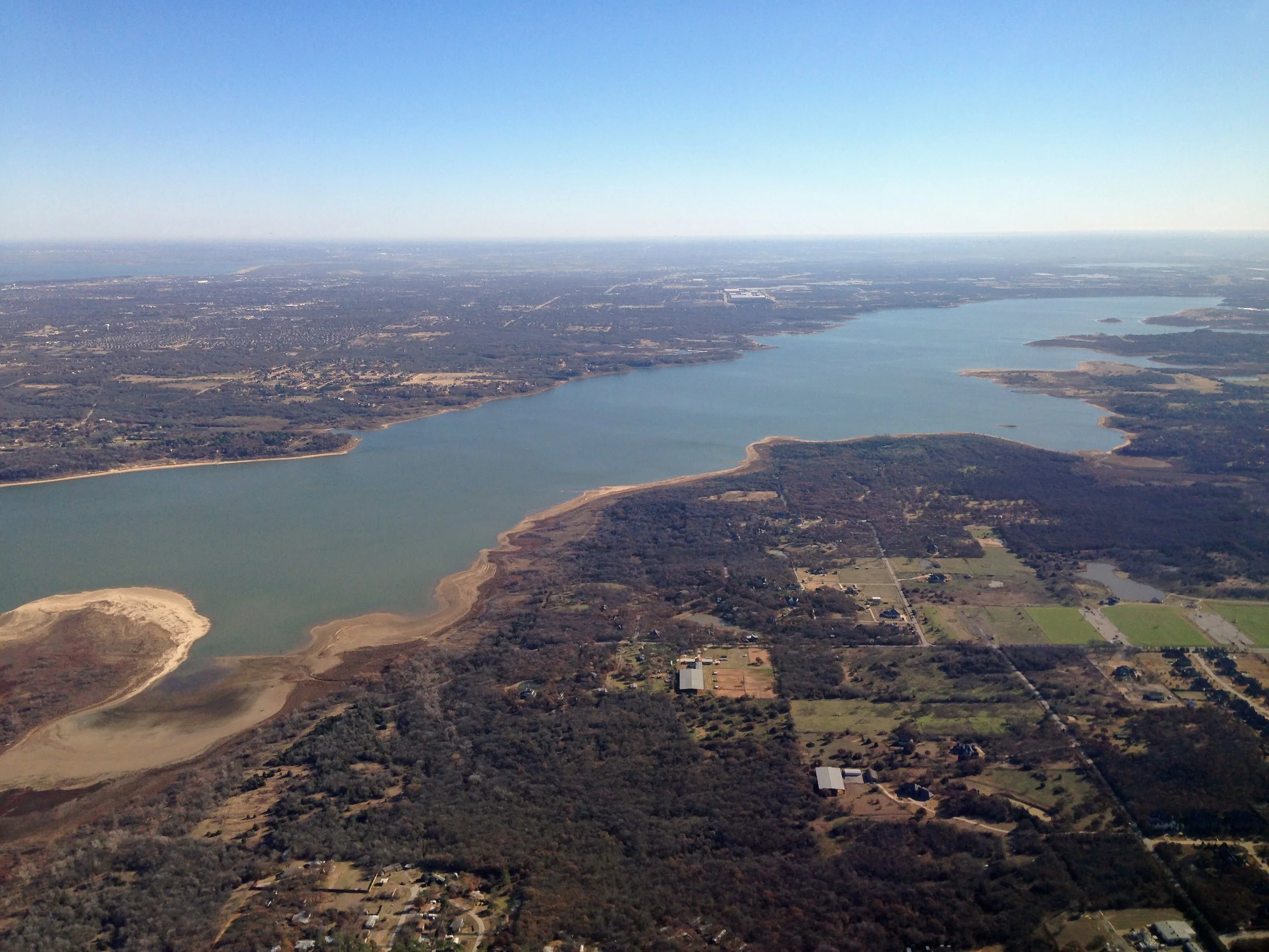 Aerial view of a large lake with surrounding landscape including forests, fields, and a few buildings, under a clear blue sky.