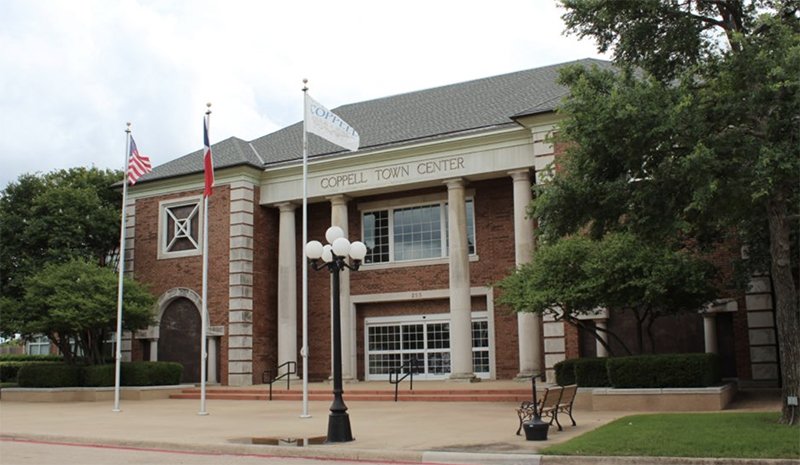 Coppell Town Center, a brick building with columns and flags, including the American flag. There is a lamppost, benches, trees, and a small lawn outside.