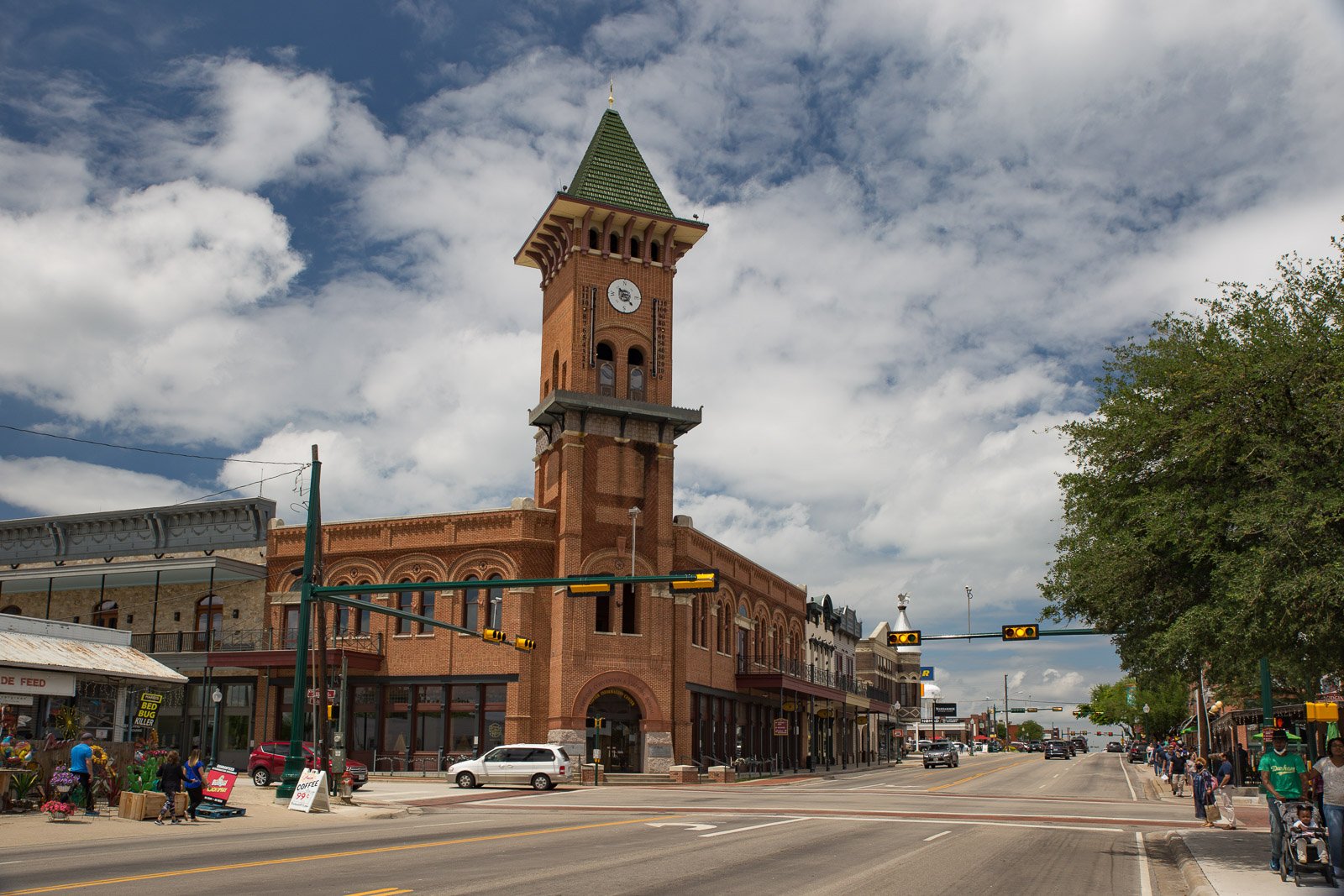A historic brick building with a clock tower on a busy street with pedestrians and cars, under a partly cloudy sky.