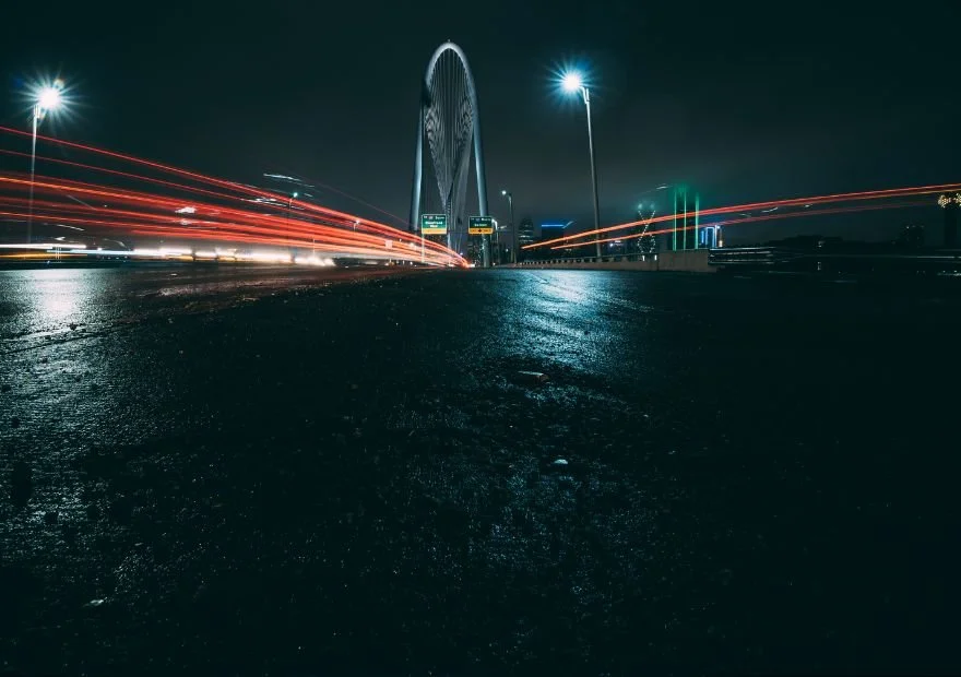 Night view of a city highway leading toward a modern, illuminated arch bridge with light trails from moving vehicles on wet asphalt.
