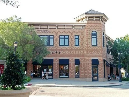A two-story brick building with retail stores on the ground floor and upper floor windows, located on a city street with trees and pedestrians.