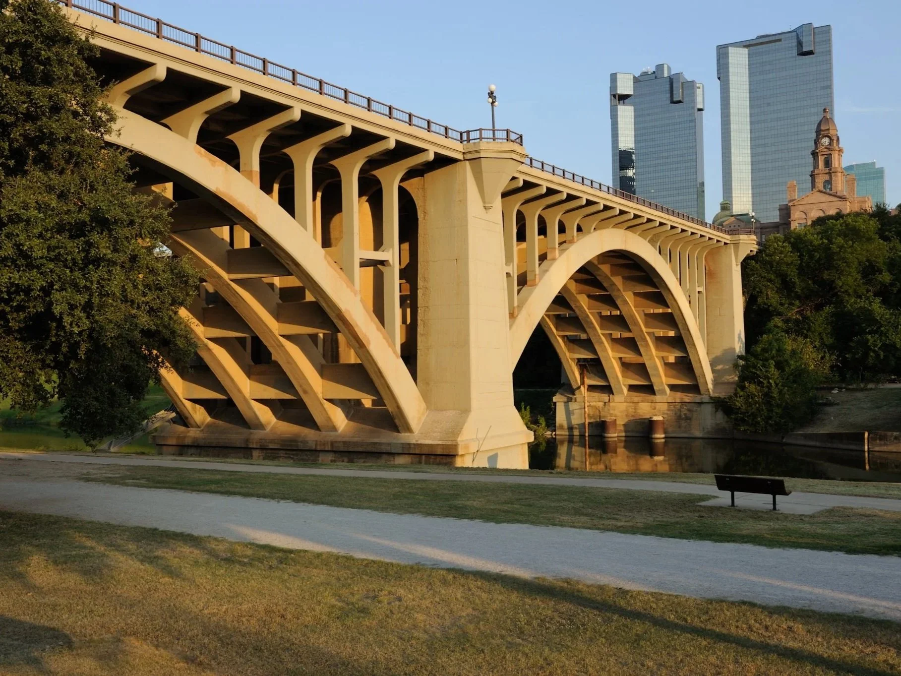 A large concrete arch bridge over a calm river, with green trees and a city skyline with tall buildings in the background, during what appears to be late afternoon or early evening just outside downtown Fort Worth, Texas