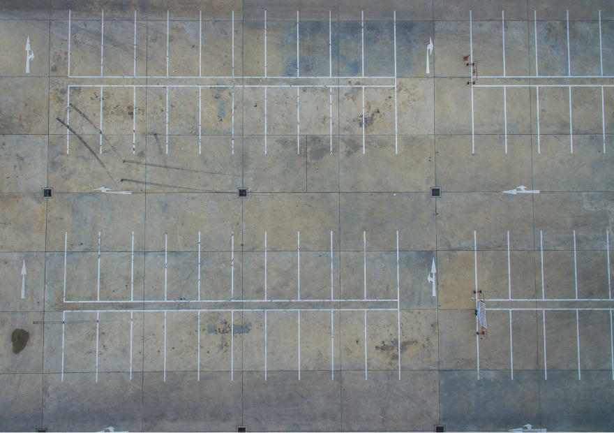 Empty parking lot with white painted parking space lines, directional arrows, and drainage grates, viewed from above.