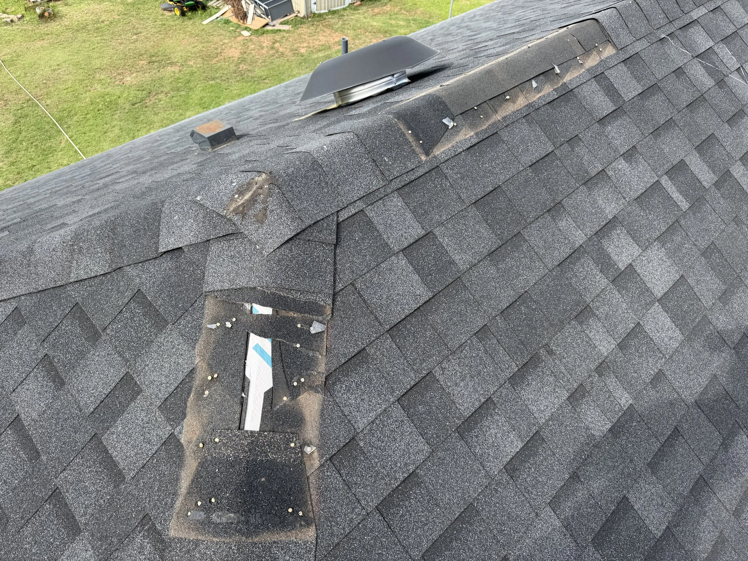 A black asphalt shingled roof with missing shingles and debris, including small rocks and pieces of roofing material, scattered on the surface. In the background, a yard with grass, a shed, and various items are visible.