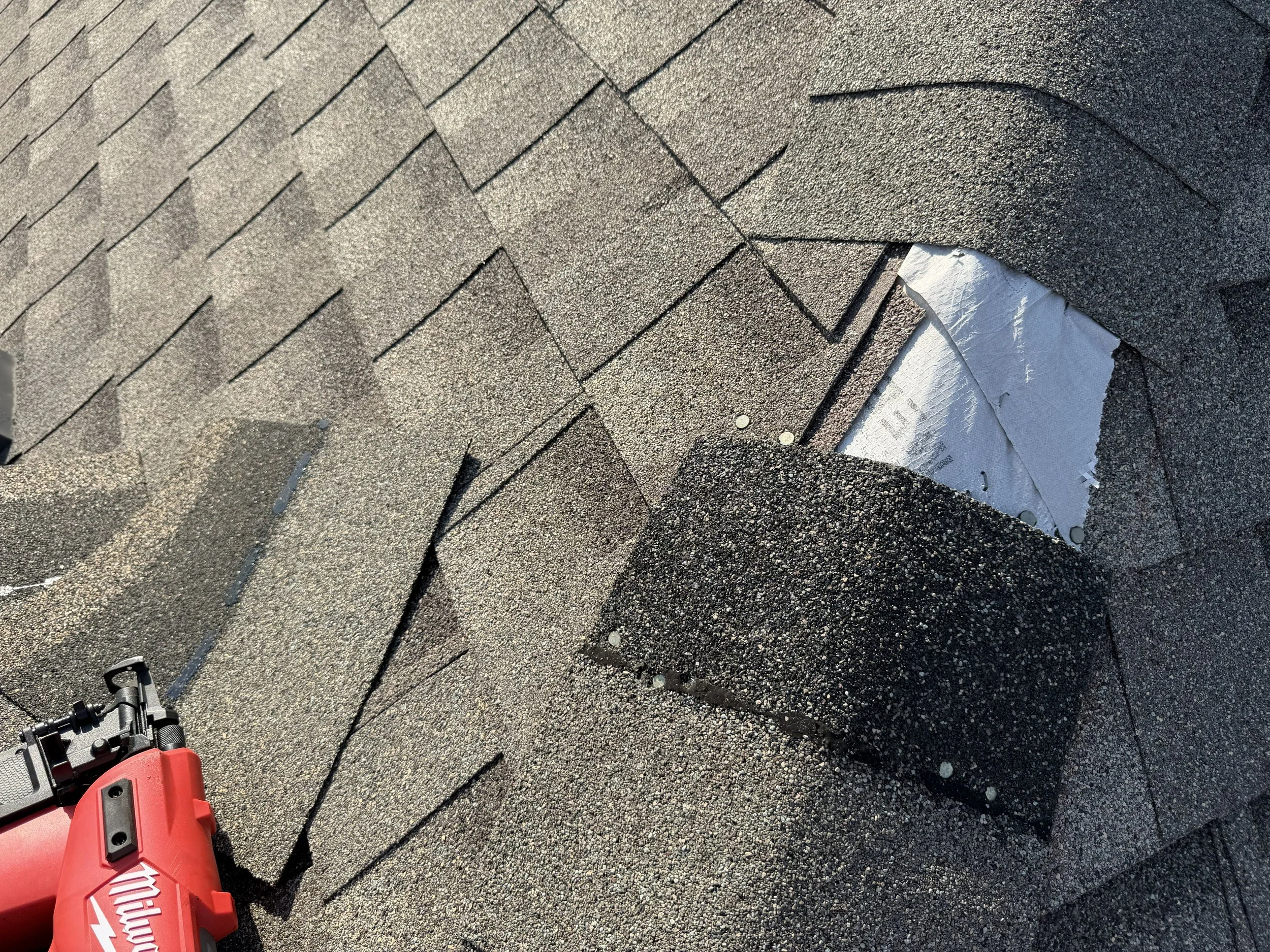 A section of a roof with asphalt shingles, some of which are being replaced. There is a power tool at the bottom left corner.