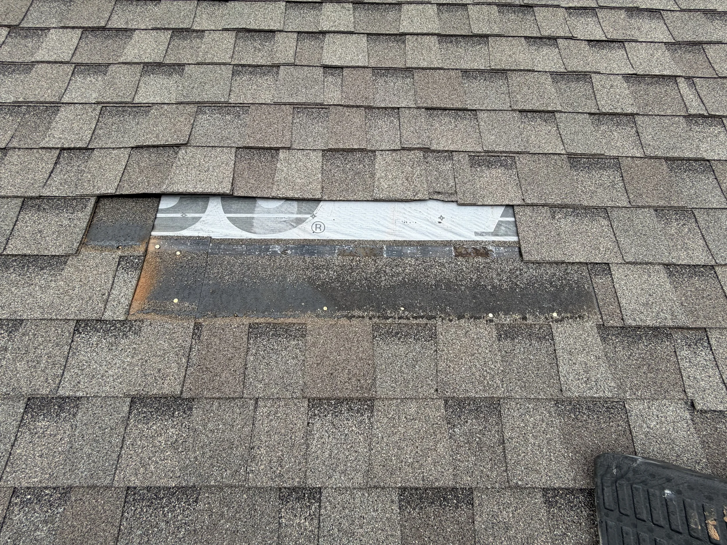 Close-up of a roof with asphalt shingles and a missing section revealing underlying roofing material.
