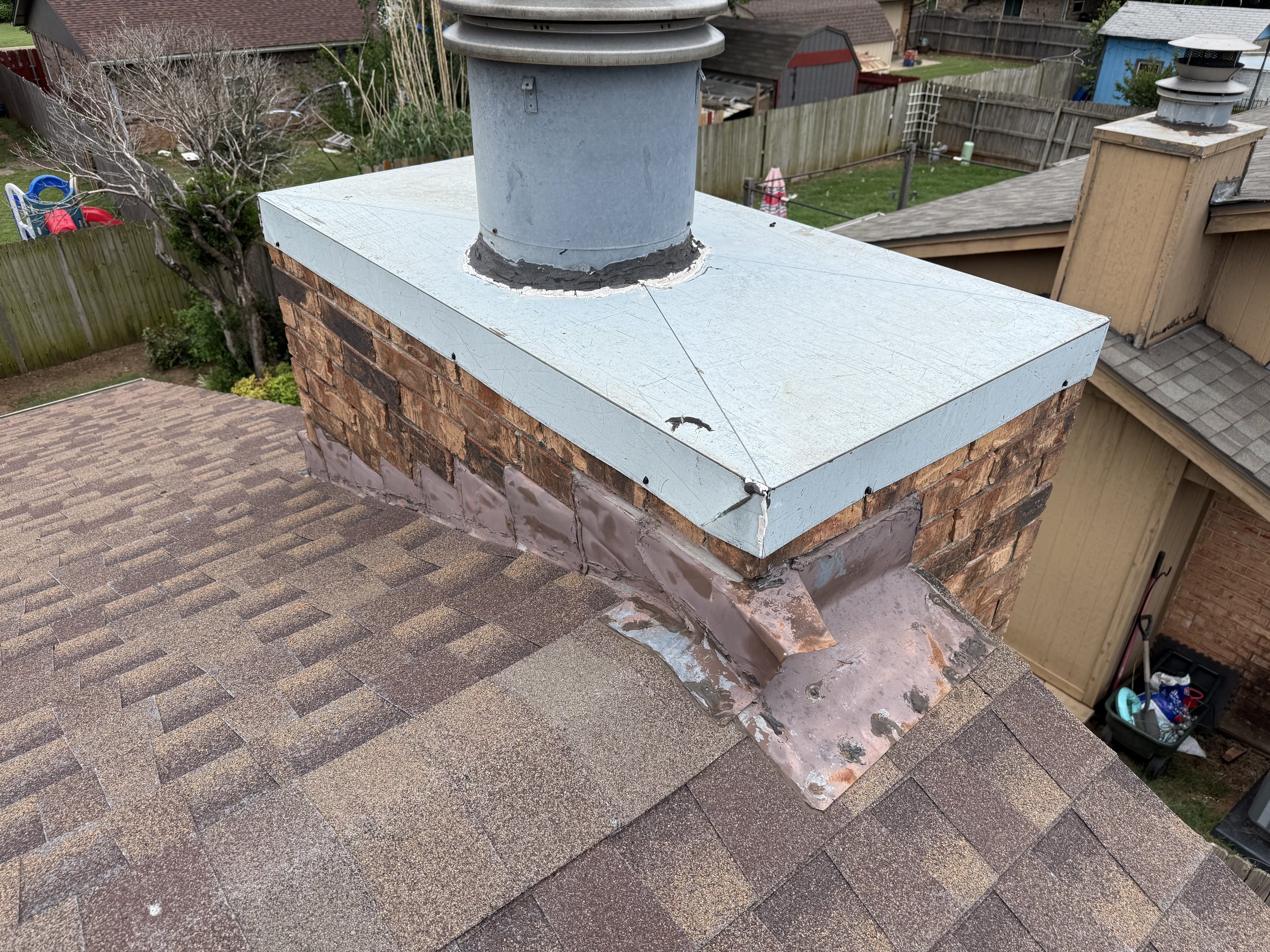 A roof vent and chimney on a residential house with visible cracks and damage around the base of the vent, overlooking a backyard with a tree, fence, and small outdoor structures.