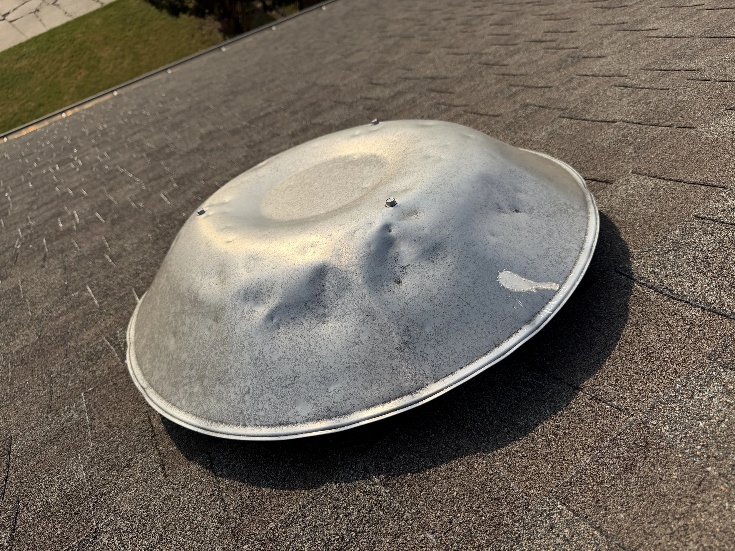 A close-up of a section of a house roof with a metal vent pipe protruding, sunlight casting shadows, and shingles visible.
