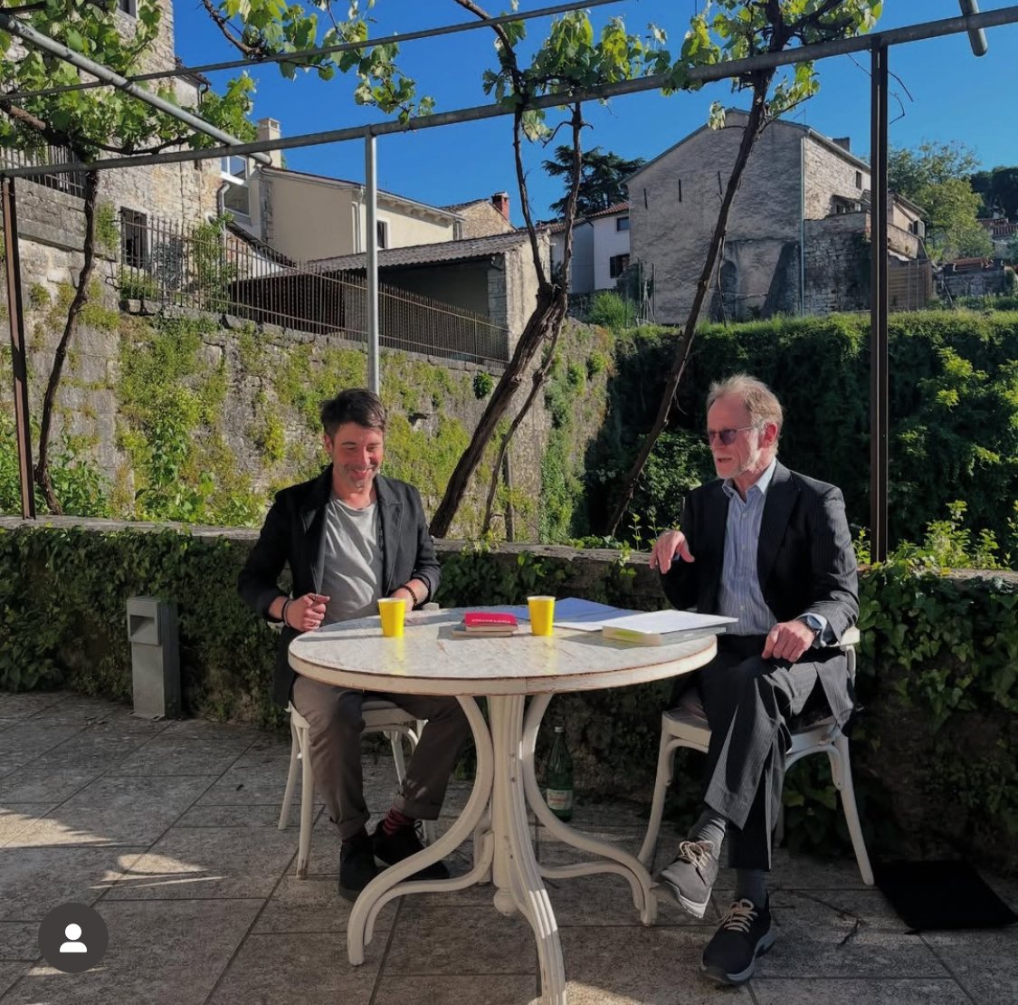 Zwei Männer sitzen an einem runden Gartentisch im Freien, lachen und sprechen. Es ist sonnig, mit Häusern und Bäumen im Hintergrund.