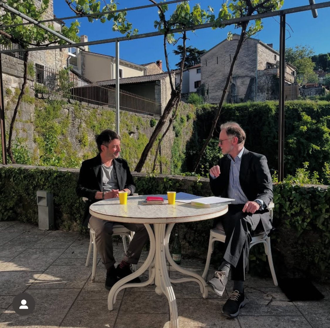 Zwei Männer sitzen an einem runden Esstisch im Freien, die Sonne scheint, mit Häusern im Hintergrund und grünen Pflanzen, während sie sprechen.