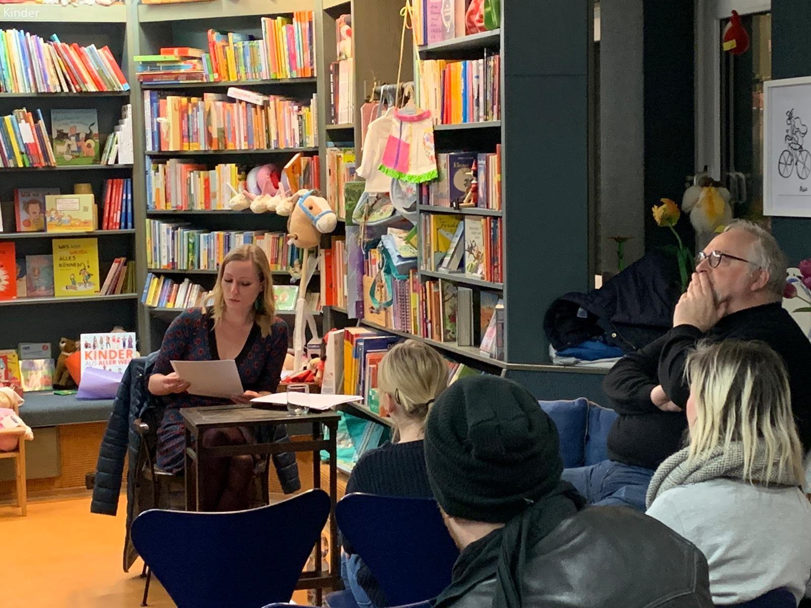 Eine Gruppe von Erwachsenen sitzt in einer Bibliothek und hört einer Frau zu, die am Tisch vorliest. Die Bibliothek ist mit bunten Büchern und Kinderartikeln gefüllt.