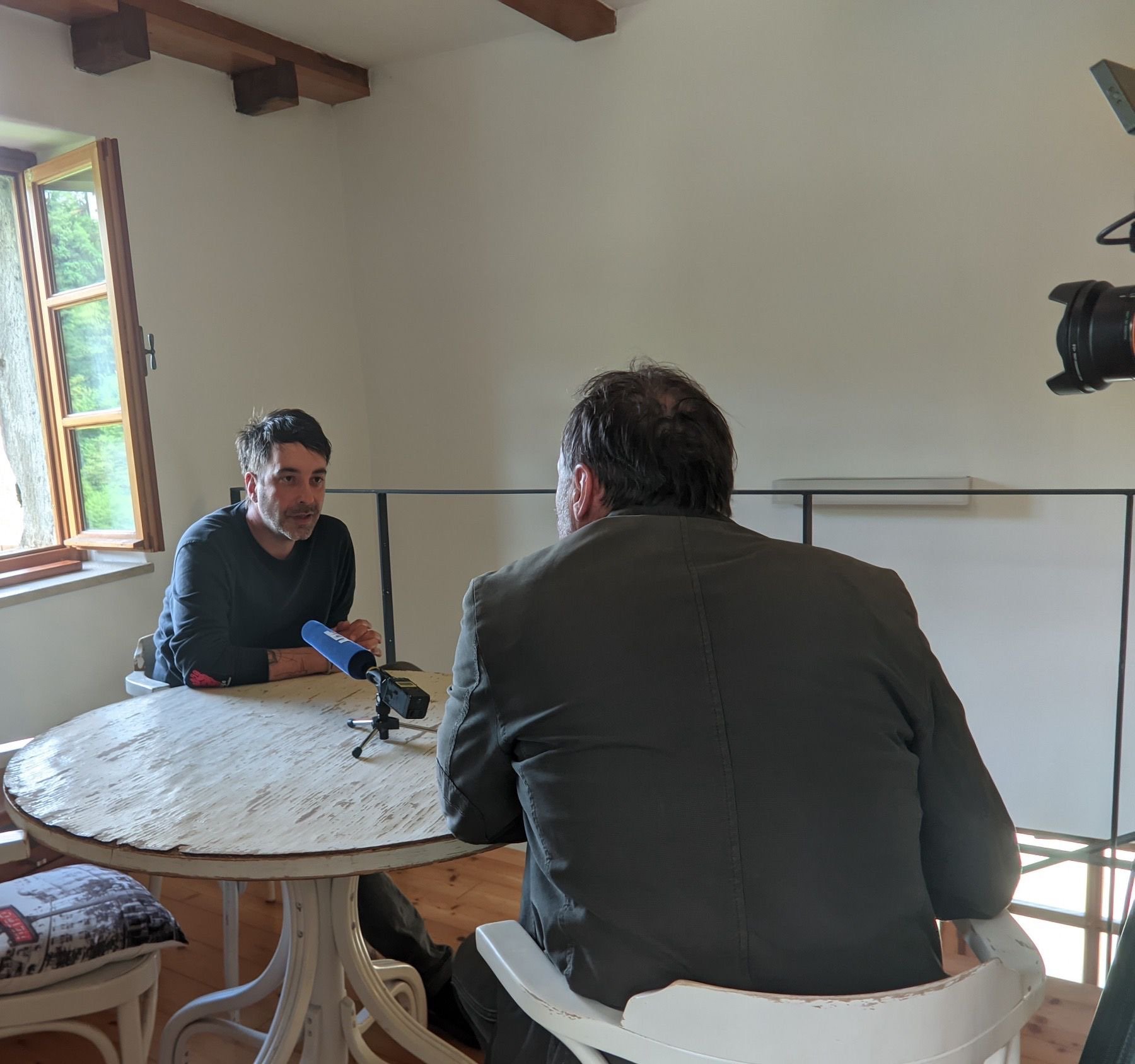 Zwei Männer im Gespräch während eines Interviews in einem hellen Raum mit einem runden Holztisch und offenen Fenster.