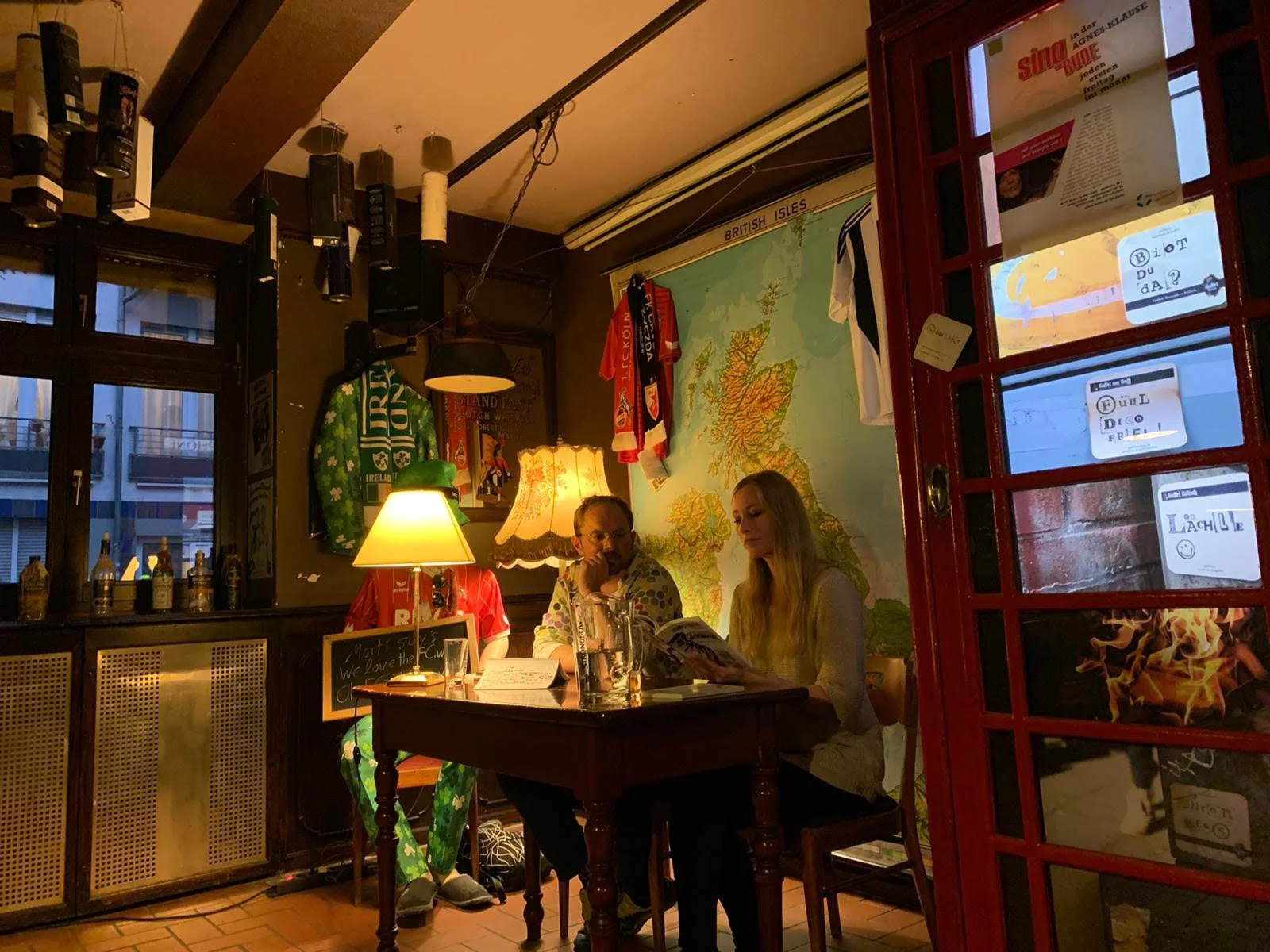 Drei Menschen sitzen an einem Tisch in einer gemütlichen Bar oder einem Pub, die Wände sind mit Karten, Kleidung und Postern dekoriert, es gibt eine Lampe mit warmem Licht auf dem Tisch.