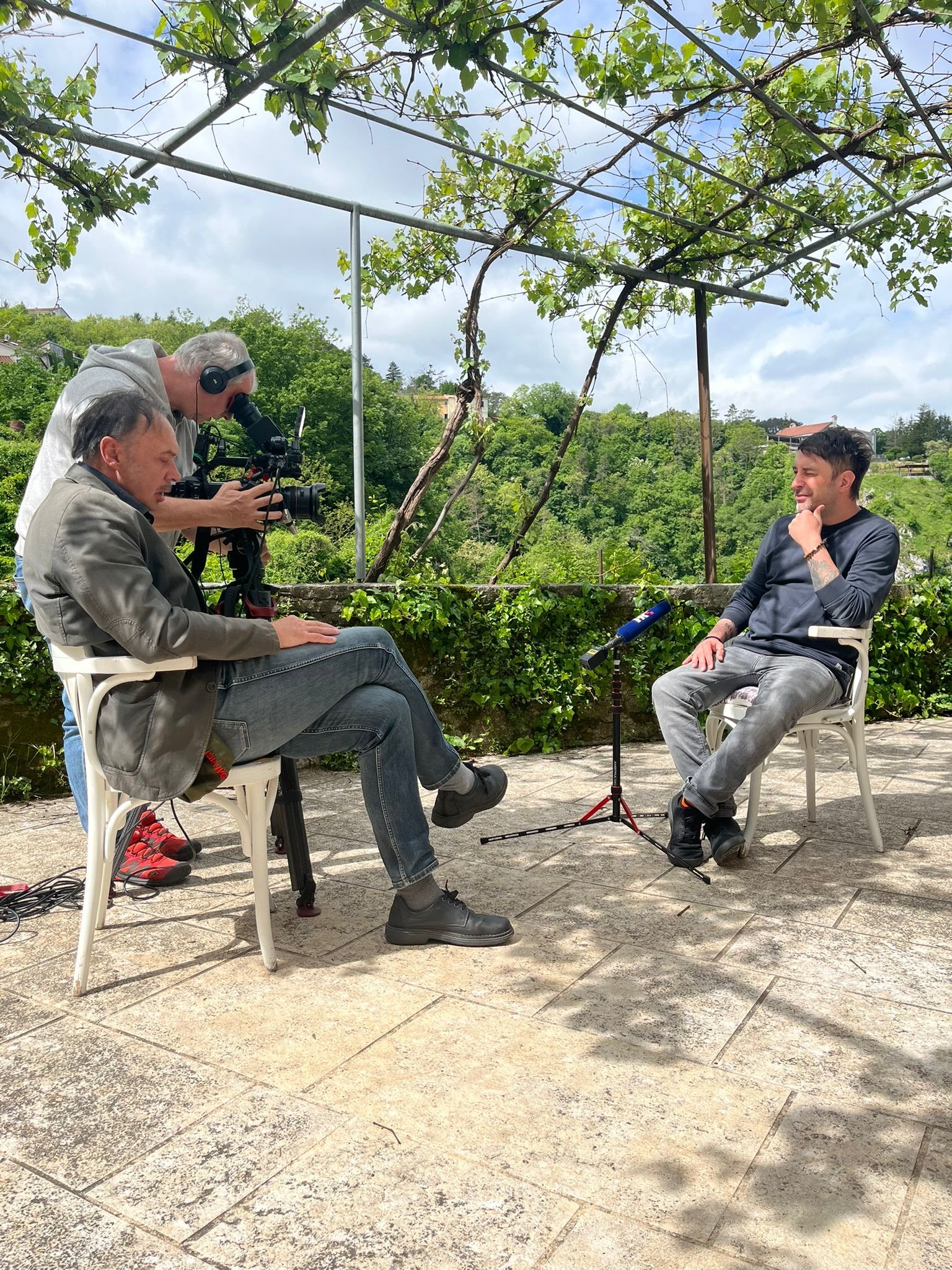 Ein Mann wird für ein Interview im Freien gefilmt, während er auf einem Stuhl sitzt und von einem Kameramann mit Kamera und einem Tonmann mit Mikrofon beobachtet wird. Im Hintergrund sind Bäume und ein bewölkter Himmel zu sehen.