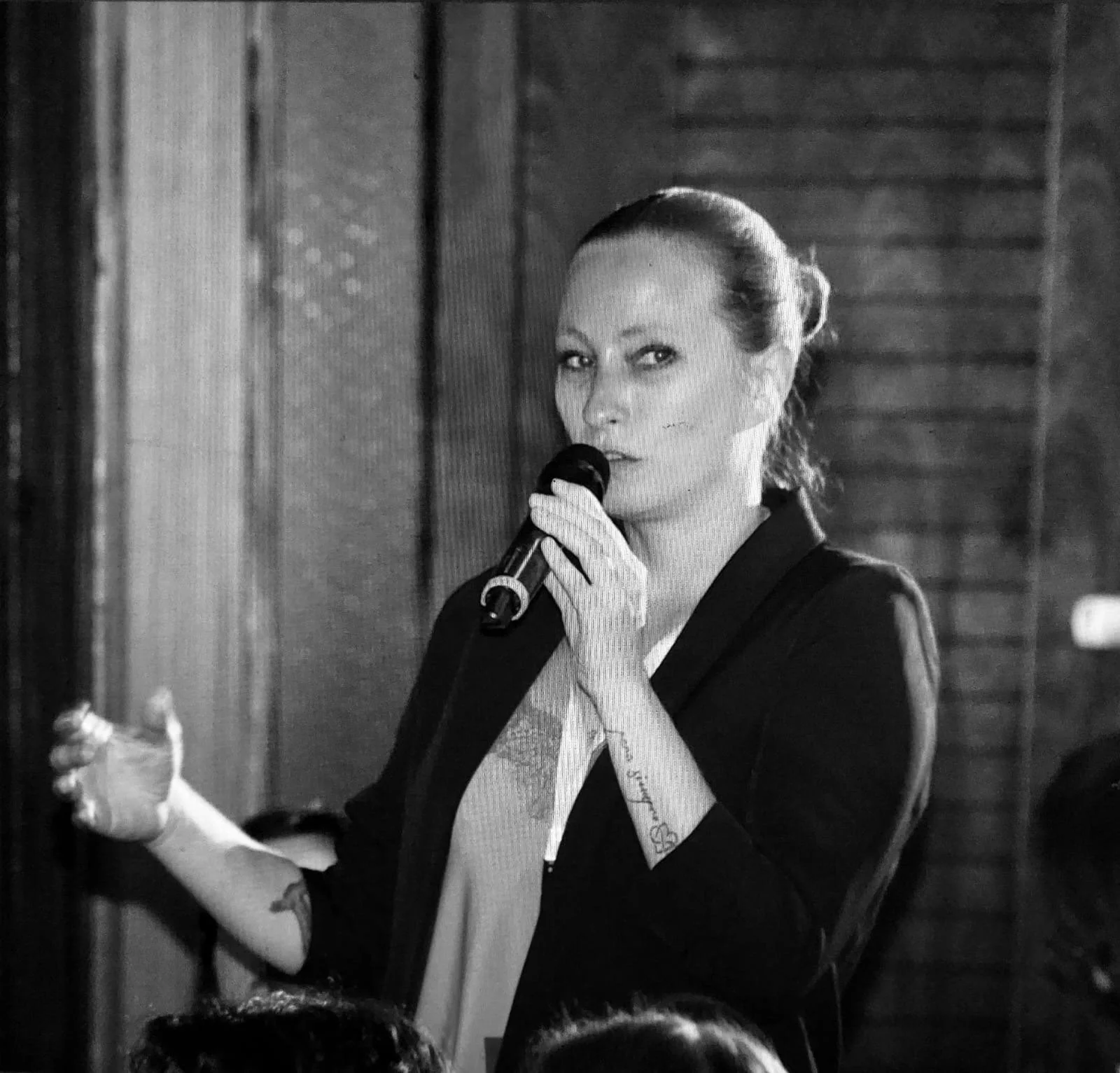 Eine Frau mit einem Mikrofon in der Hand spricht vor einem Publikum in einem Raum mit holzvertäfelten Wänden.