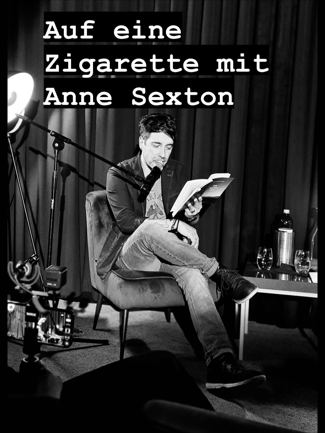 Schwarz-Weiß-Foto eines Mannes, der auf einem Stuhl sitzt, in eine Lesebrille schaut und ein Buch liest, mit einem Mikrofon vor ihm und einem Hintergrund aus Vorhängen. Text auf Bild: 'Auf eine Zigarette mit Anne Sexton'.