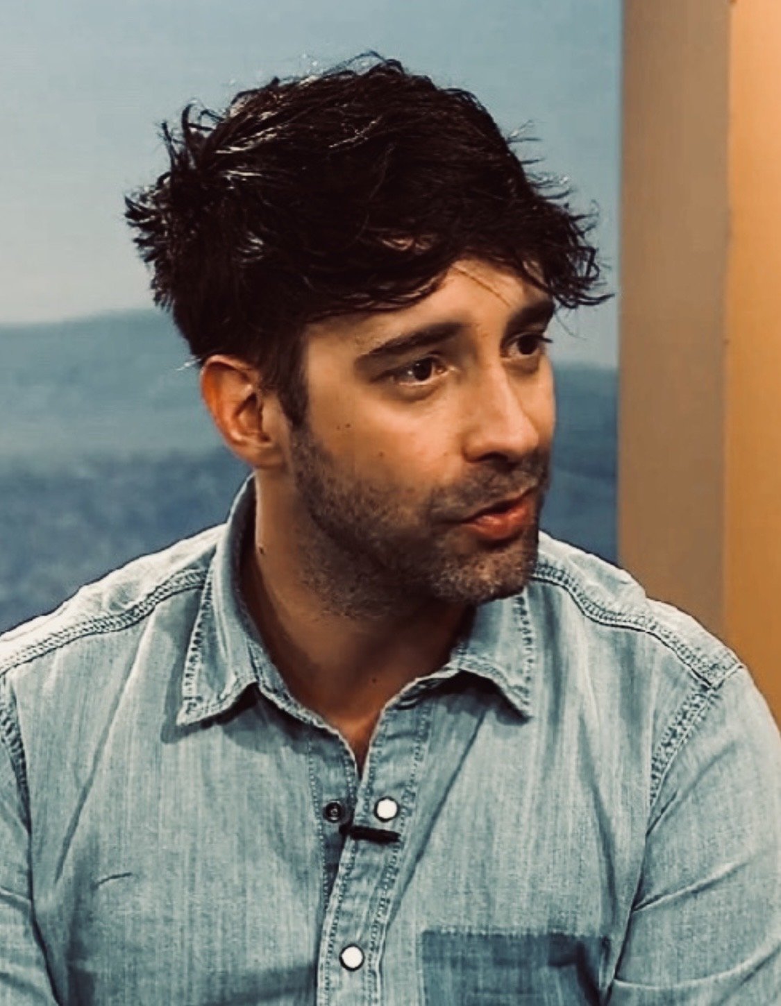 Ein Mann mit dunklen, gewellten Haaren und Bart trägt eine hellblaue Jeansjacke und sitzt an einem Ort mit blauer und orangefarbener Wand. Er schaut nach rechts.