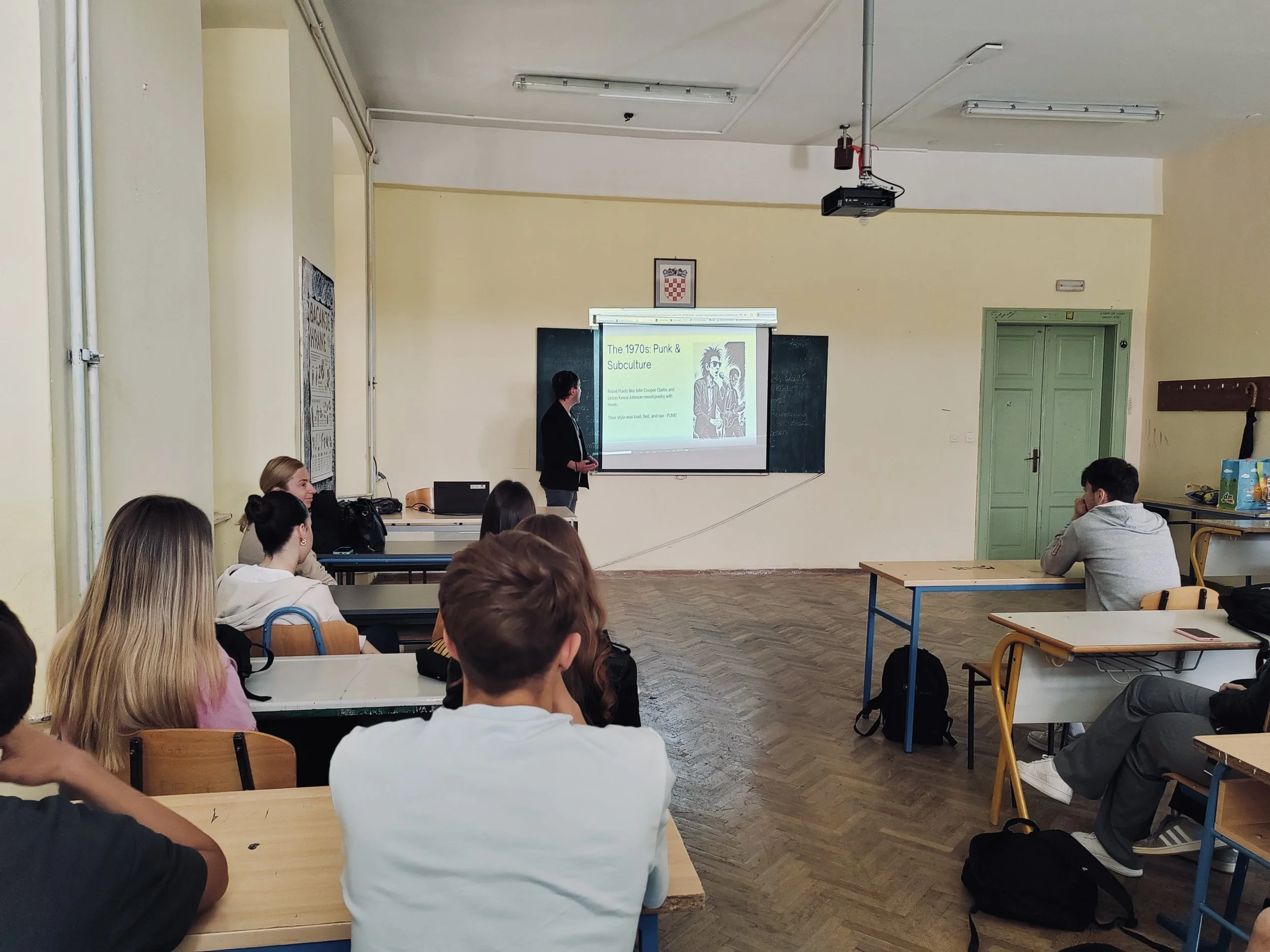 Ein Klassenraum mit Schülern, die einem Vortrag zuhören, an der Tafel ist eine Präsentation über die 1970er Jahre Punk- und Subkultur zu sehen.