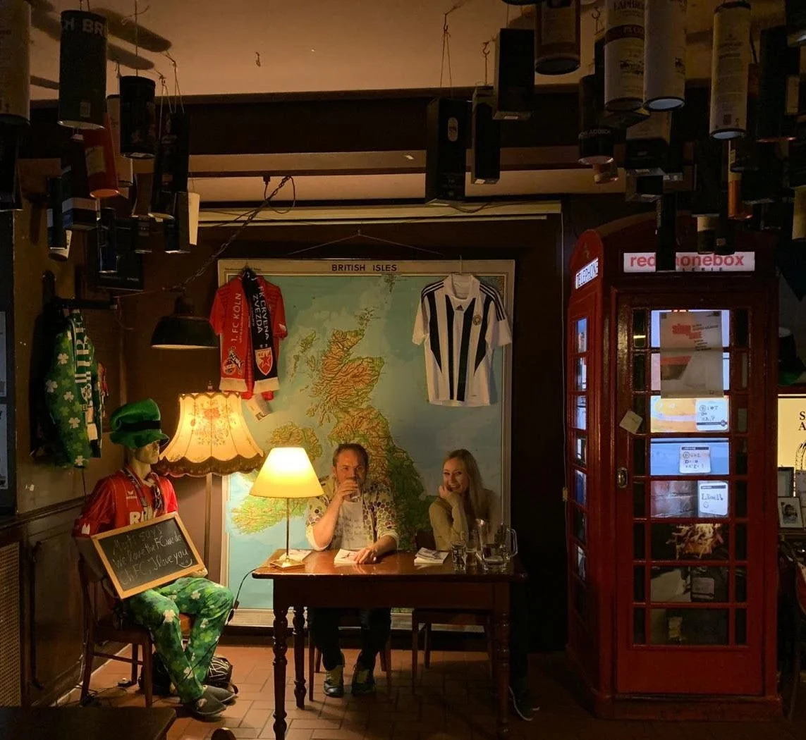 Ein Pub mit Wandkarte des Britischen Inseln, einer Personendisplay mit Fußballtrikots und einer roten Telefonzelle. Zwei Leute sitzen an einem Tisch, eine Person trägt ein rotes Fußballtrikot, die andere unterhält sich. Es gibt eine Stehlampe, einen alten Lampenschirm und einen Sprechblasen-Prop mit einem Gruß. Die Szene ist warm beleuchtet und vermittelt eine gemütliche Atmosphäre.