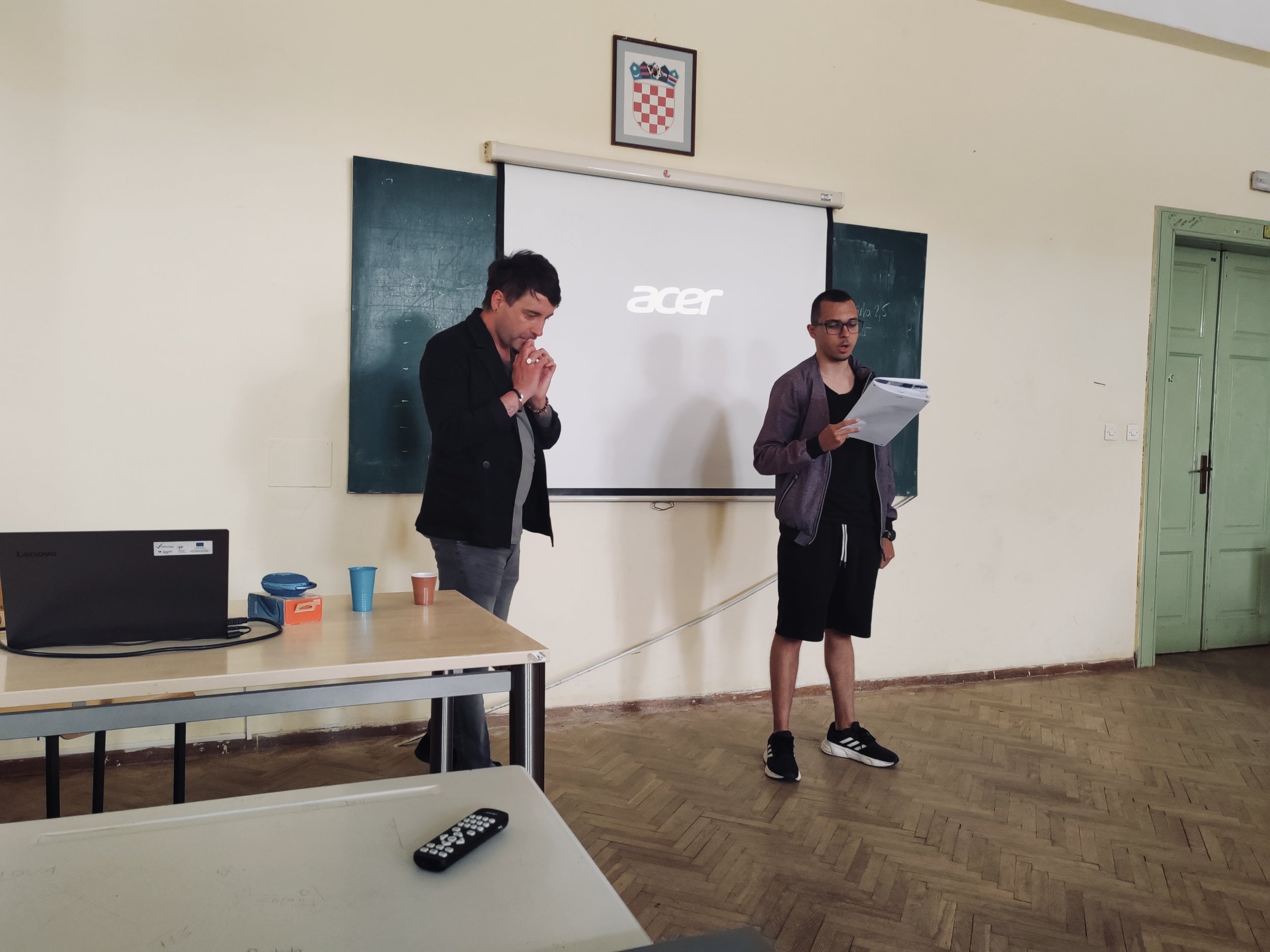 Zwei junge Männer stehen in einem Klassenraum vor einer Tafel, einer liest etwas von einem Stapel Papier, der andere steht mit verschränkten Armen. Auf dem Tisch liegen eine Laptop, Tassen und ein Fernbedienung.