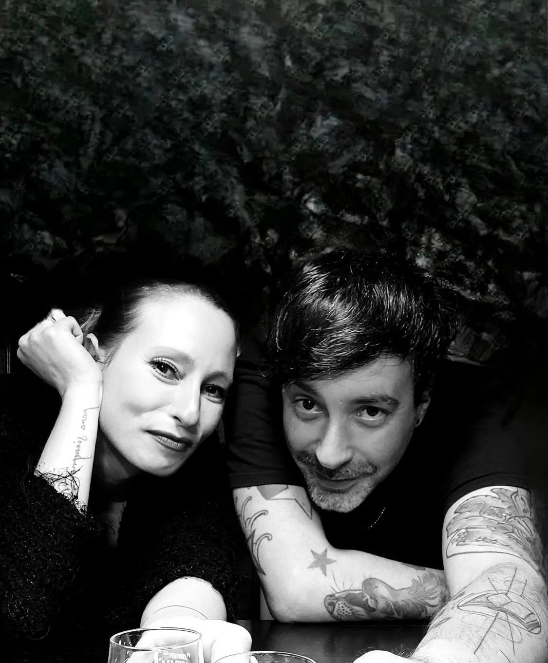 Zwei Menschen liegen auf einem Tisch, schauen in die Kamera, tätowiert, eine Frau mit dunklem Haar und ein Mann mit dunkler gestylter Frisur, im Hintergrund eine dunkle, strukturierte Wand.