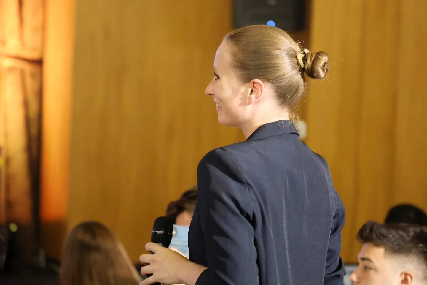 Eine junge Frau in einem dunklen Blazer steht seitlich im Raum und hält ein Mikrofon. Sie lächelt. Hinter ihr sind mehrere Menschen, von denen einige Masken tragen, und die Wand ist aus Holz.