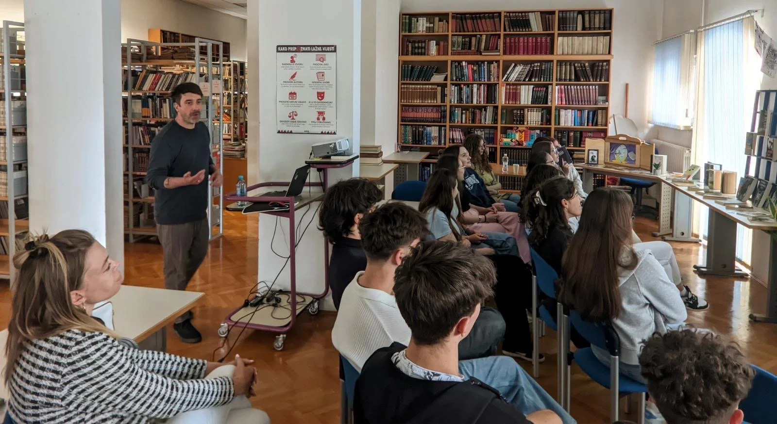 Ein Mann hält einen Vortrag vor einer Gruppe junger Menschen in einer Bibliothek, Regal mit Büchern im Hintergrund, einige sitzen an Tischen, andere auf Stühlen, bei Tageslicht.