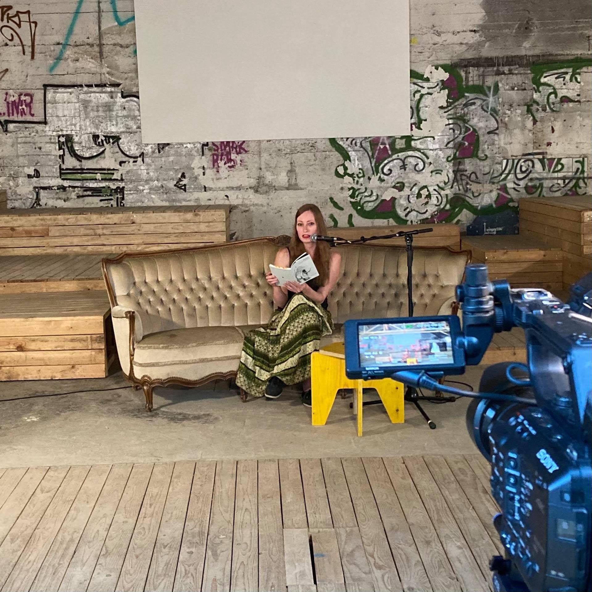 Eine Frau sitzt auf einem antiken Sofa, liest aus einem Buch vor einem Mikrofon, während eine Kamera sie filmt. Hintergrund: eine Graffiti-Wand mit bunten Designs und Schriftzügen. Im Vordergrund ein Kameramonitor.