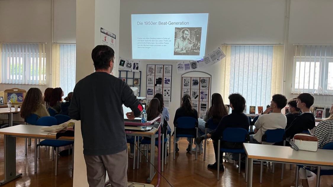 Ein Lehrer zeigt einen Vortrag über die Beat-Generation der 1950er Jahre vor einer Gruppe von Schülern in einem Klassenraum, mit einem Projektor an der Wand.