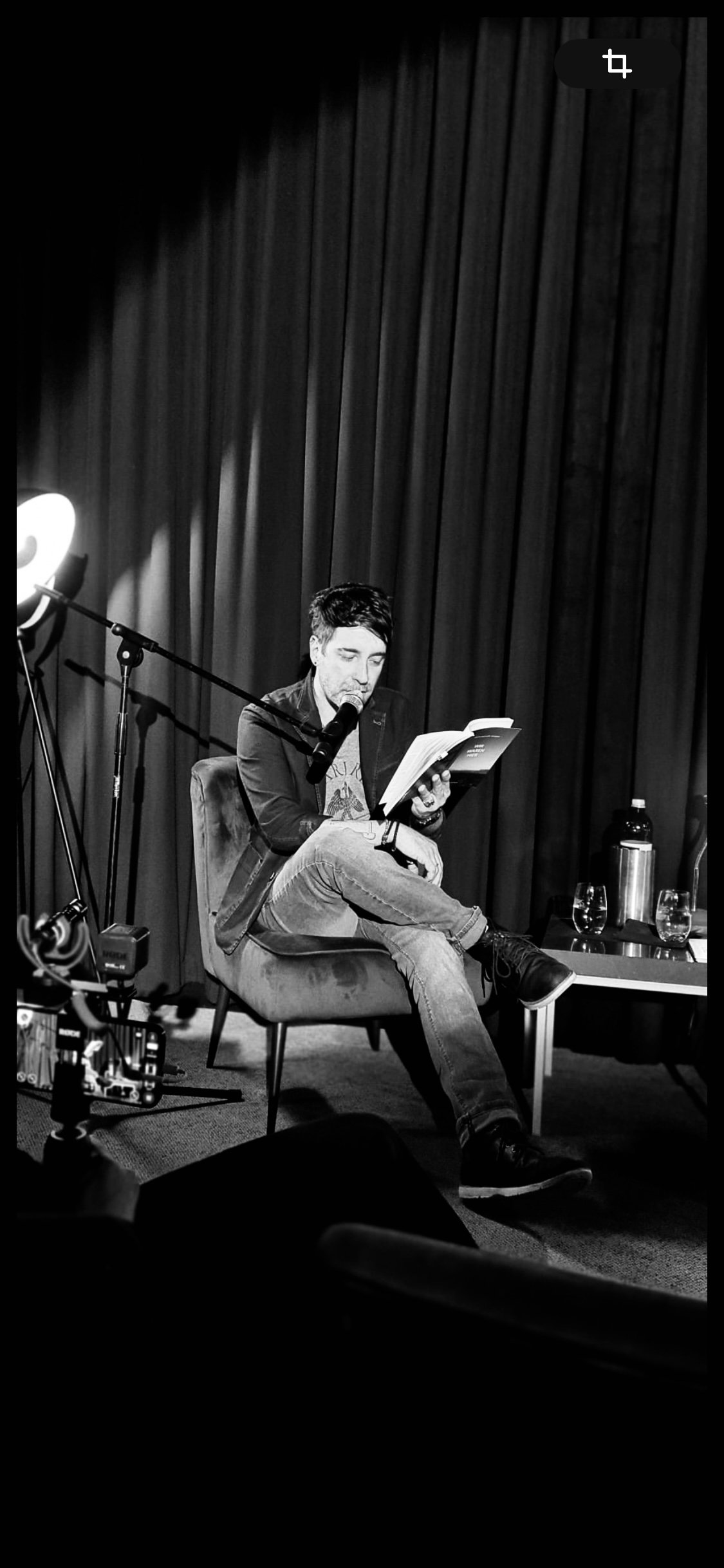 Mann liest in einem schwarzen und weißen Bild auf einem Stuhl sitzend aus einem Buch, mit einer Mikrofon vor ihm, im Hintergrund ein Vorhang. Es gibt Gegenstände auf einem Tisch, darunter eine Wasserflasche, Gläser und einen Thermobecher.