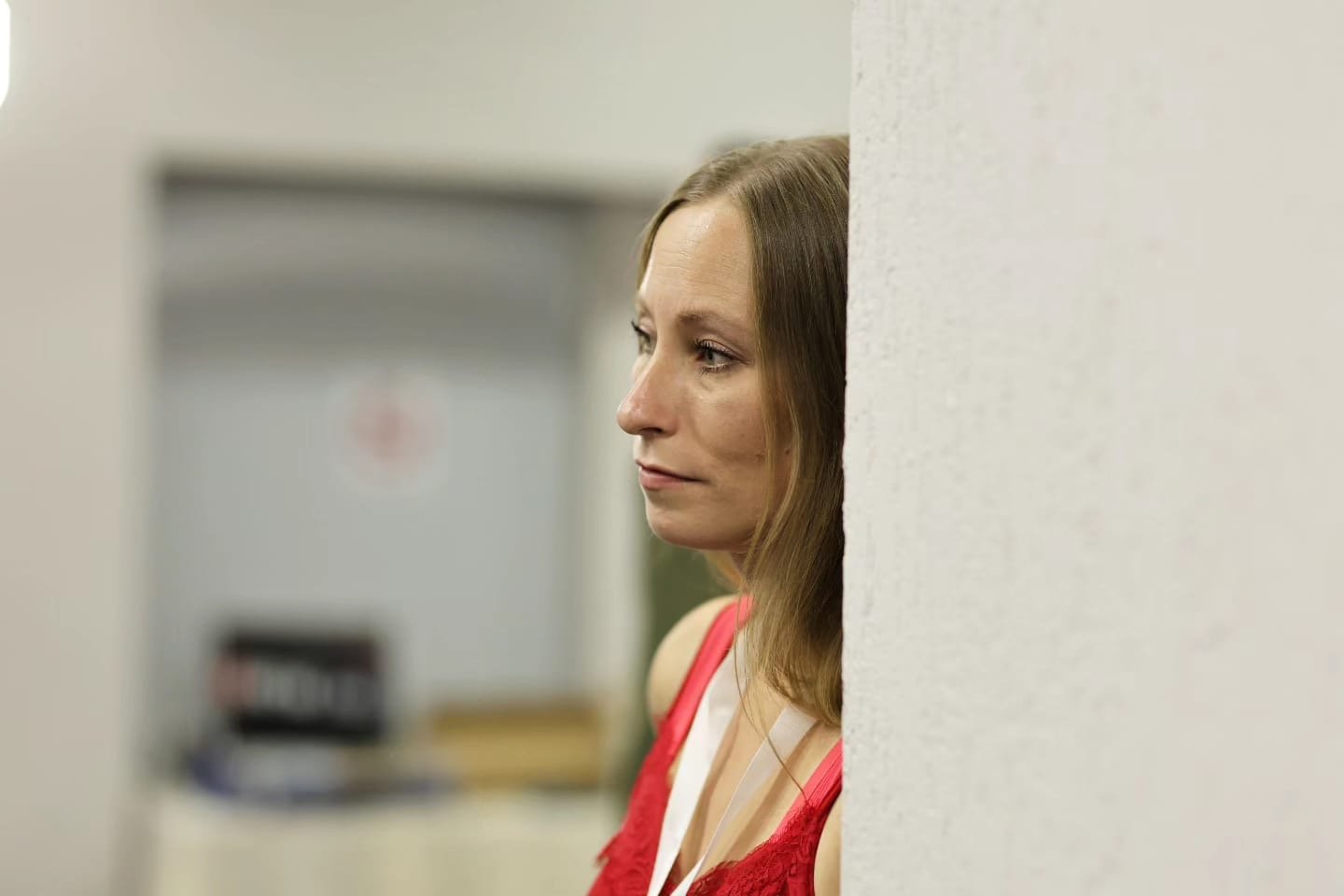 Eine Frau mit braunen Haaren, die in einem roten Top vor einer weißen Wand steht, blickt nach links, während sie an der Wand lehnt.