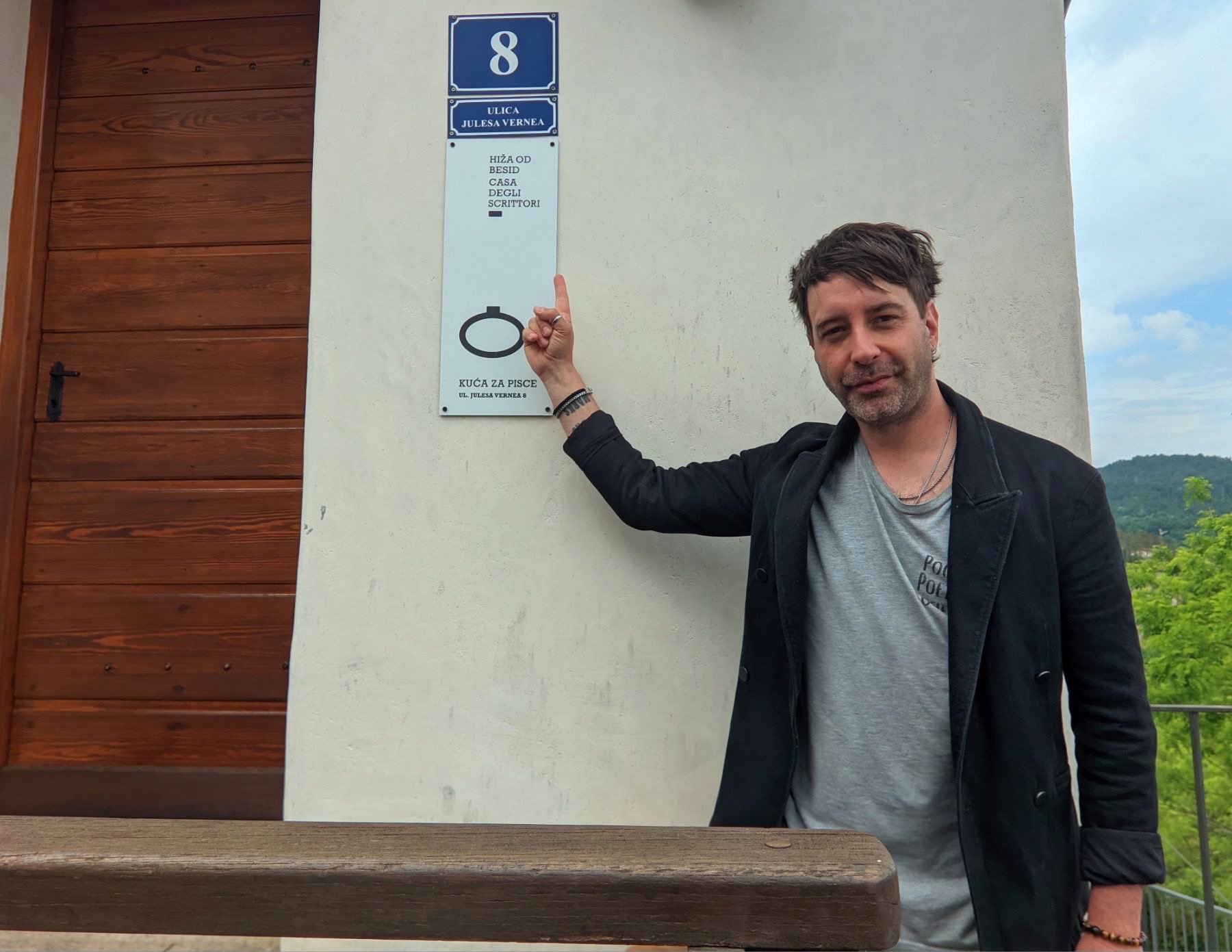 Ein Mann steht neben einer Hausnummerntafel, die die Adresse 8, Juliusa Vernea zeigt. Er zeigt auf die Tafel und lächelt.