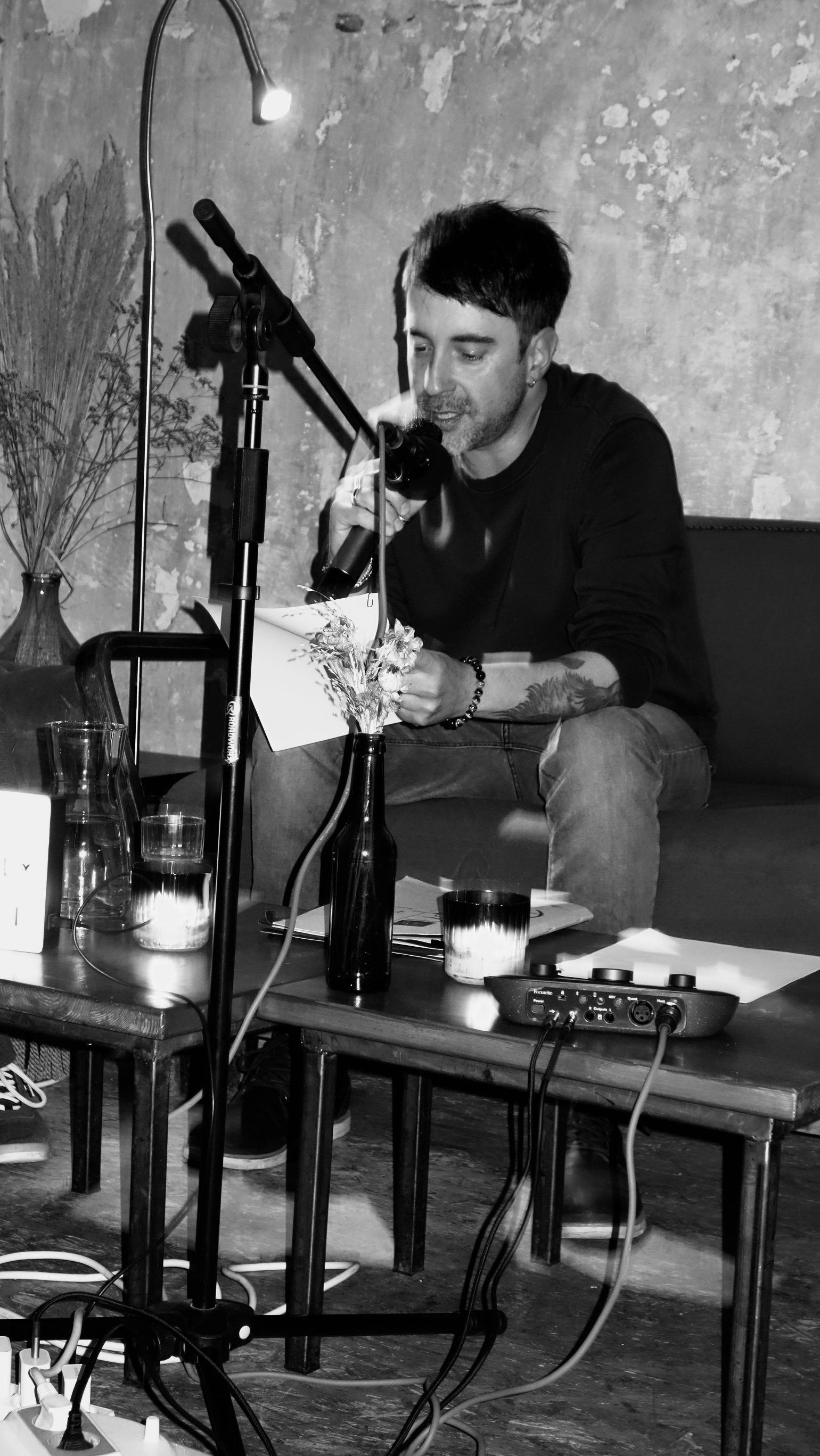 Ein Mann sitzt auf einem Sofa, liest ein Blatt mit Blumen darauf und spricht in ein Mikrofon, im Hintergrund eine Wand mit texturierter Oberfläche.