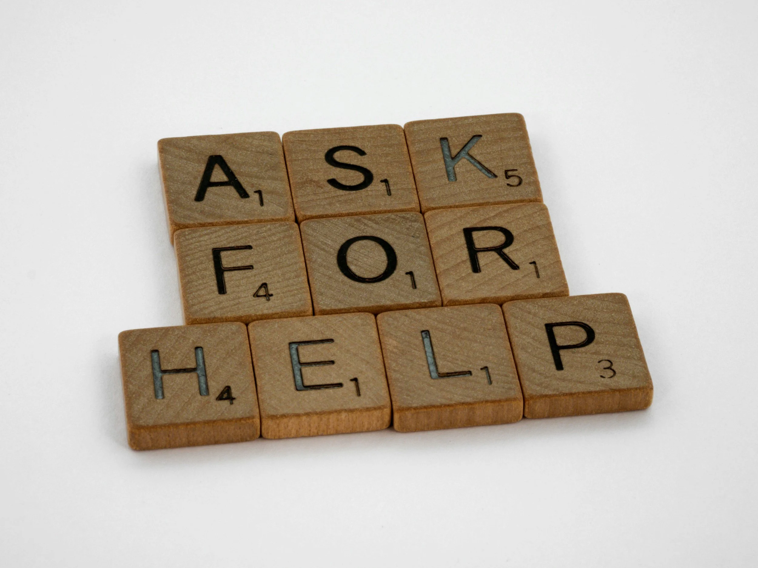 Wooden Scrabble tiles spelling out the phrase "ASK FOR HELP" on a white background.