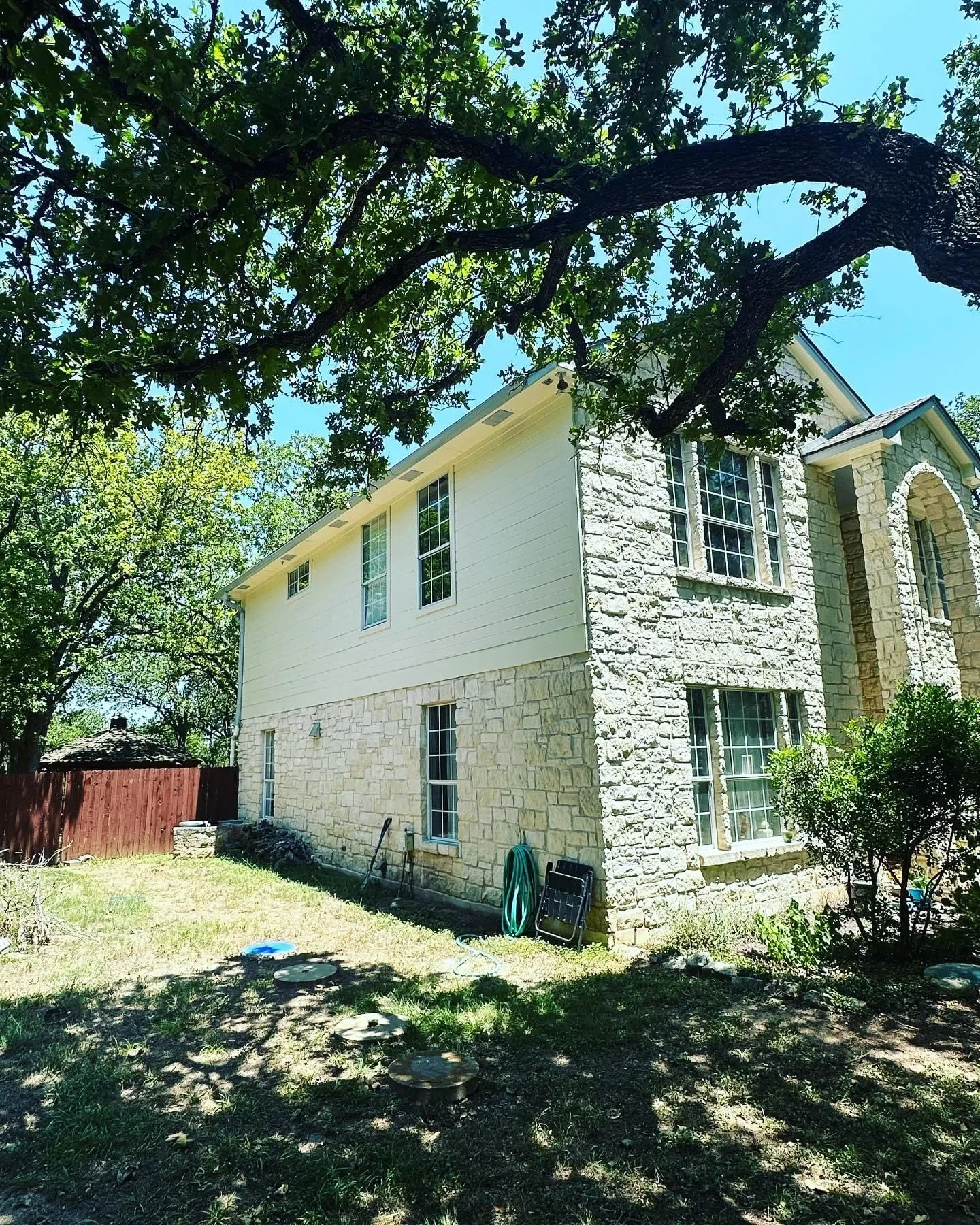 Side view of a two-story house with stone and white siding exterior, surrounded by trees and greenery, with some outdoor gardening tools and hose on the grass.