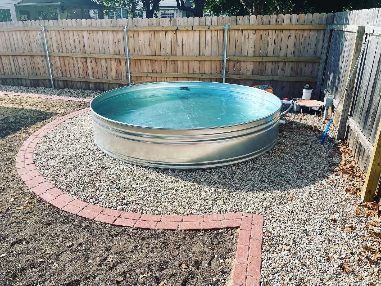 A round metal hot tub filled with water in a backyard enclosed by a wooden fence, with a gravel and brick-lined ground and some outdoor equipment in the background.