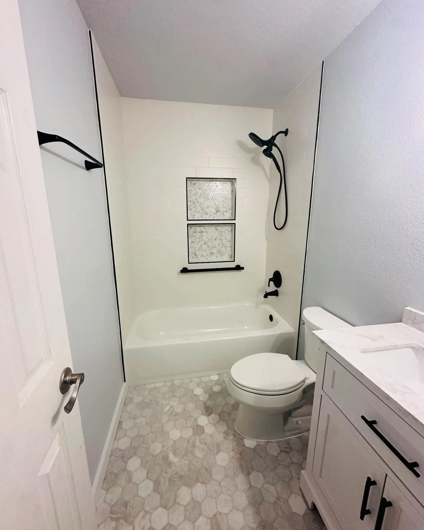 Bathroom with a white bathtub, black shower fixtures, toilet, and a white vanity with black handles. The floor has hexagonal tiles, and there is a small window with patterned glass in the shower area.