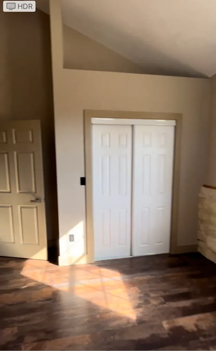 A room with hardwood flooring, a double closet door, and a partially open door, with sunlight casting a shadow on the floor.