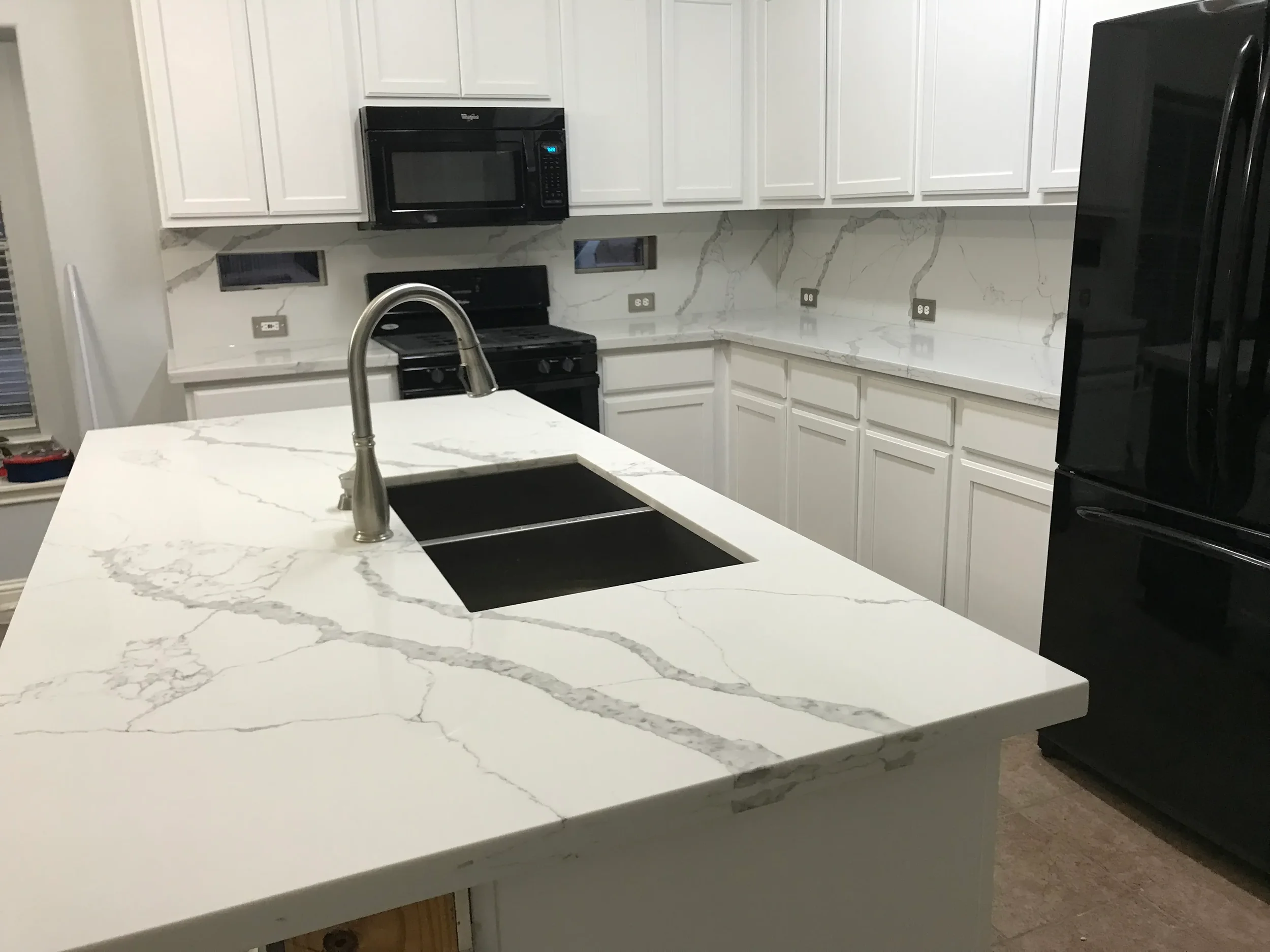 Kitchen with white cabinetry, marble countertops, black appliances including a microwave, stove, and refrigerator, and a large kitchen island with a black sink and a stainless steel faucet.
