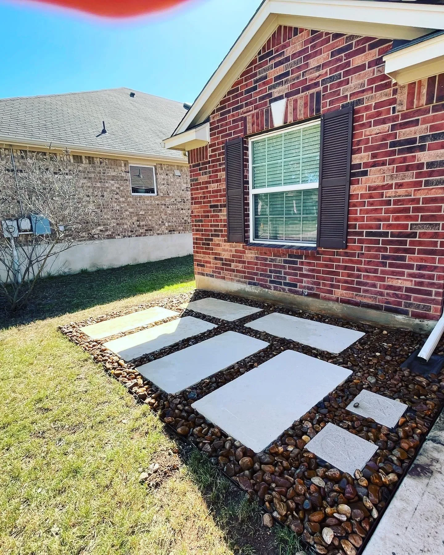 Backyard with a red brick house, black shutters, and a window. There is a small garden bed with large stepping stones and small rocks around them, adjacent to a grassy lawn.