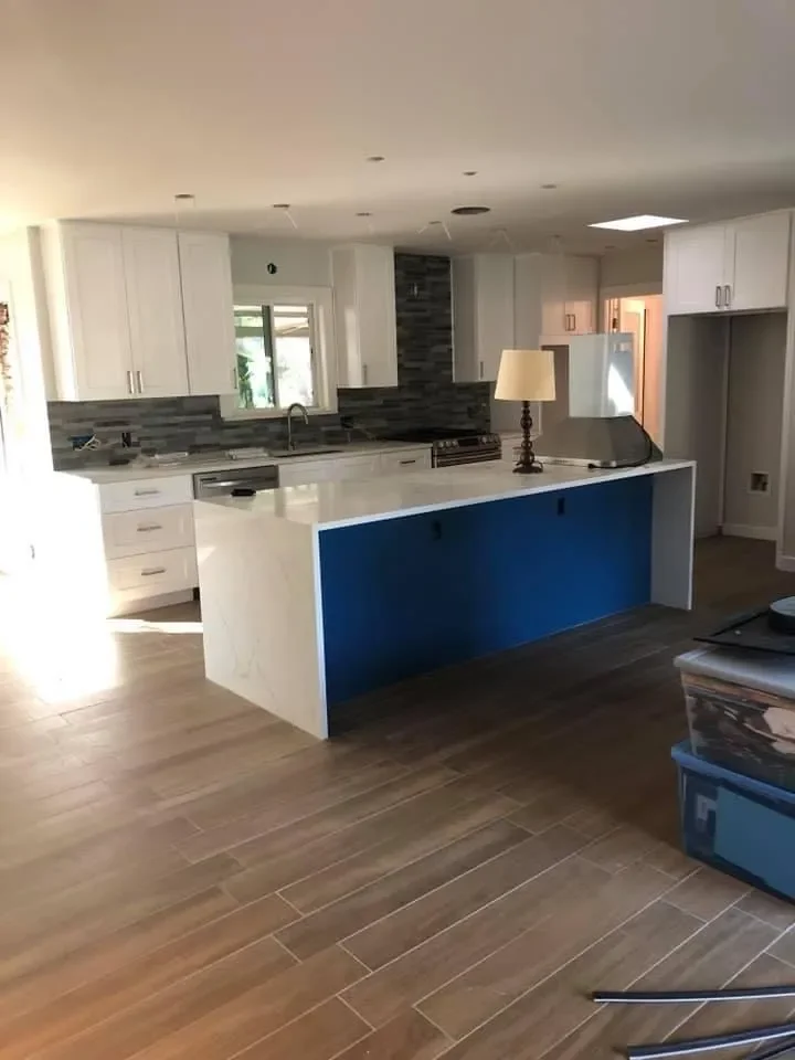 Modern kitchen with white cabinets, a large white island with a blue side panel, wooden flooring, and a dark gray backsplash. There is a small window above the sink and a lamp on the island.
