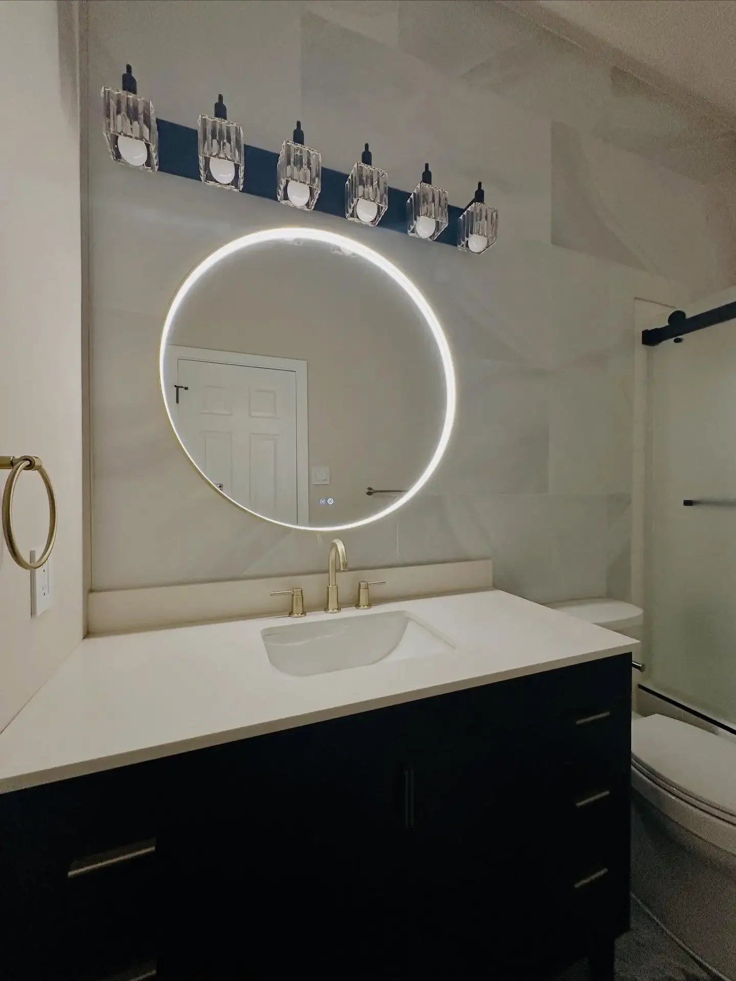 Modern bathroom with a circular LED mirror, black vanity with white countertop, gold fixtures, and a wall-mounted light fixture with multiple glass shades.