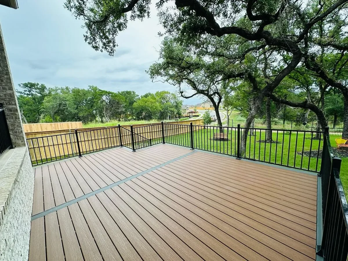 View of a backyard deck with brown wooden planks, black metal railing, trees, green grass, and a cloudy sky in the background.
