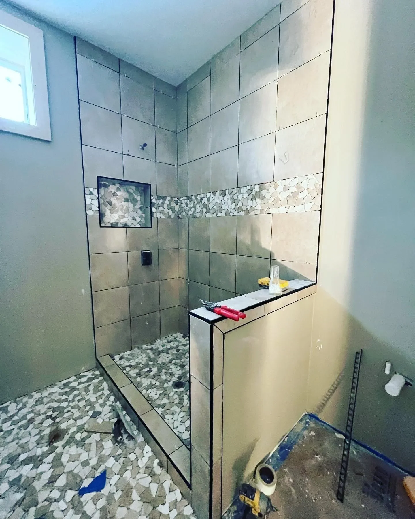 Bathroom under construction featuring a tiled shower area with large beige tiles, a decorative pebble strip, a small niche, and various construction tools and materials.