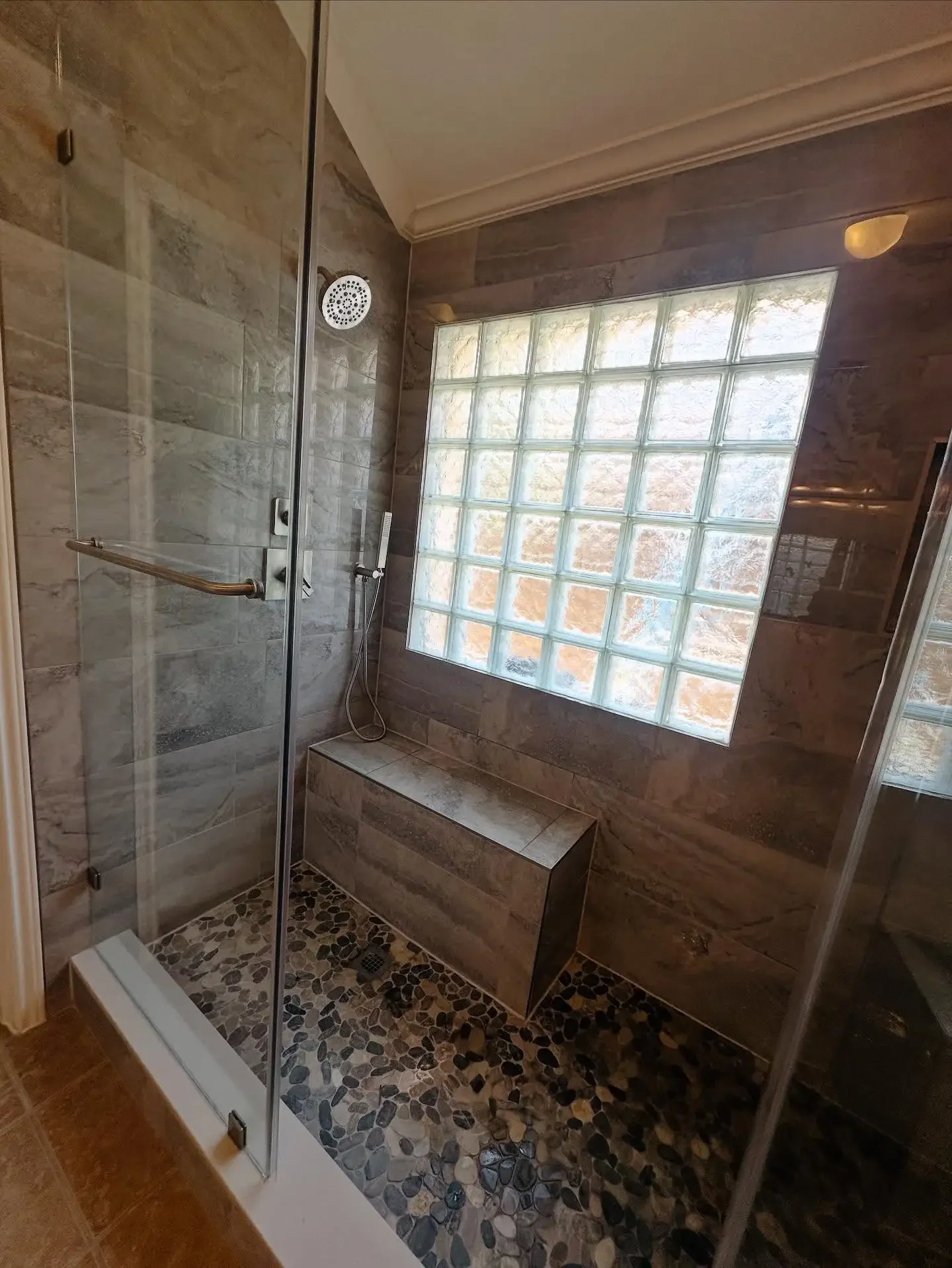 A bathroom shower area with glass walls, a pebble stone floor, a built-in bench, a window made of glass blocks, and shower fixtures.