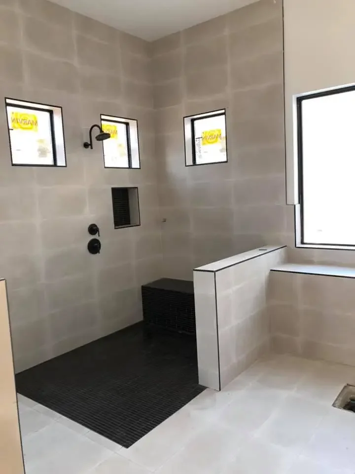 A modern, minimalistic bathroom shower area with three small high-set windows, a black showerhead, a built-in wall niche, a black bench, and a partial wall separating the shower from the bathtub, all with neutral-colored tiles.
