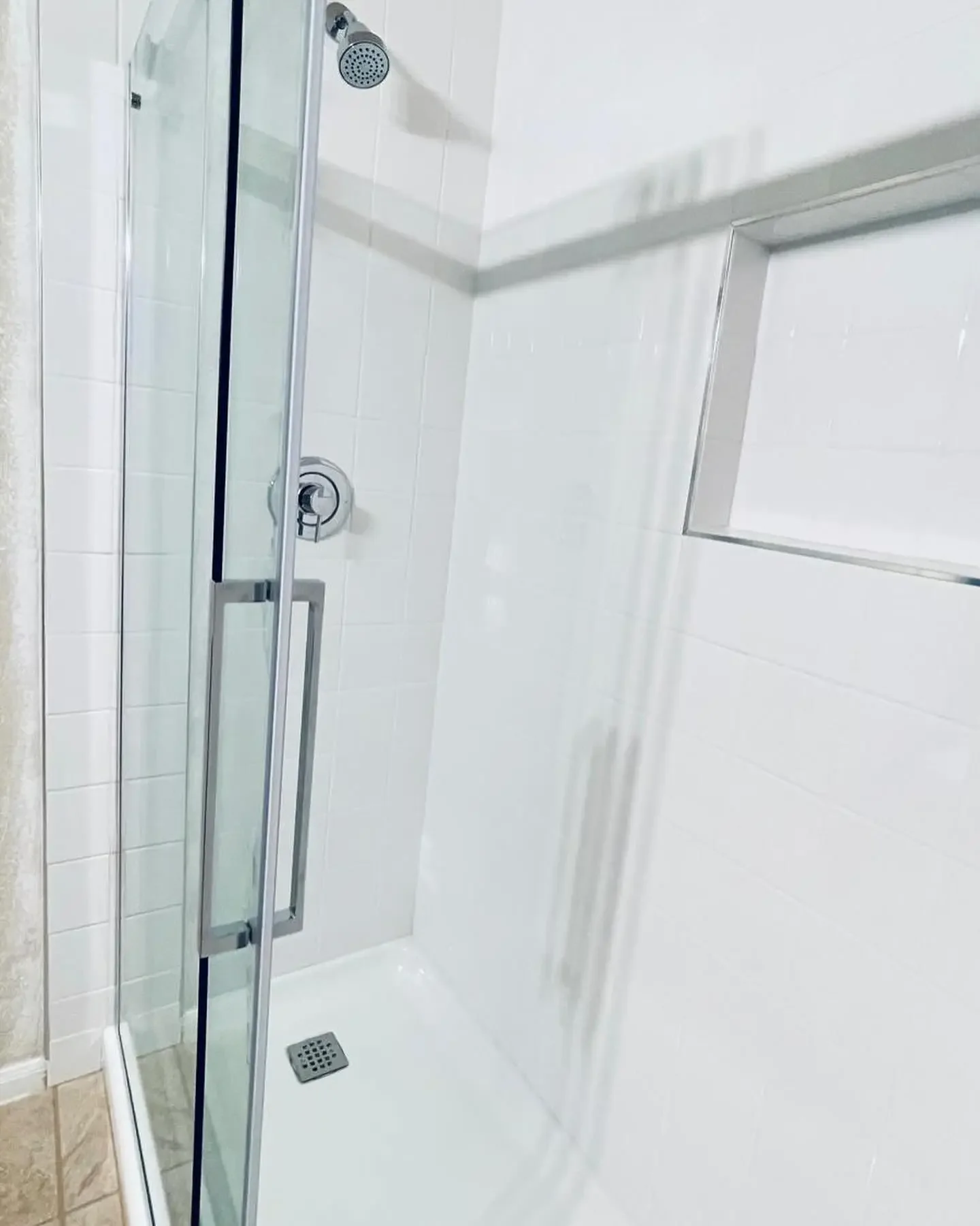 Empty glass shower enclosure with a metal handle, showerhead, and drain on white tiled walls and floor.