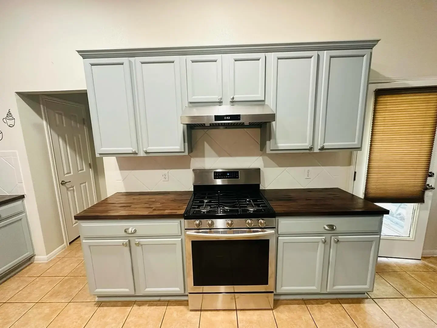 Kitchen with white cabinets, dark wood countertops, stainless steel oven and range hood, tan tiled floor, door with window and closed beige blinds.