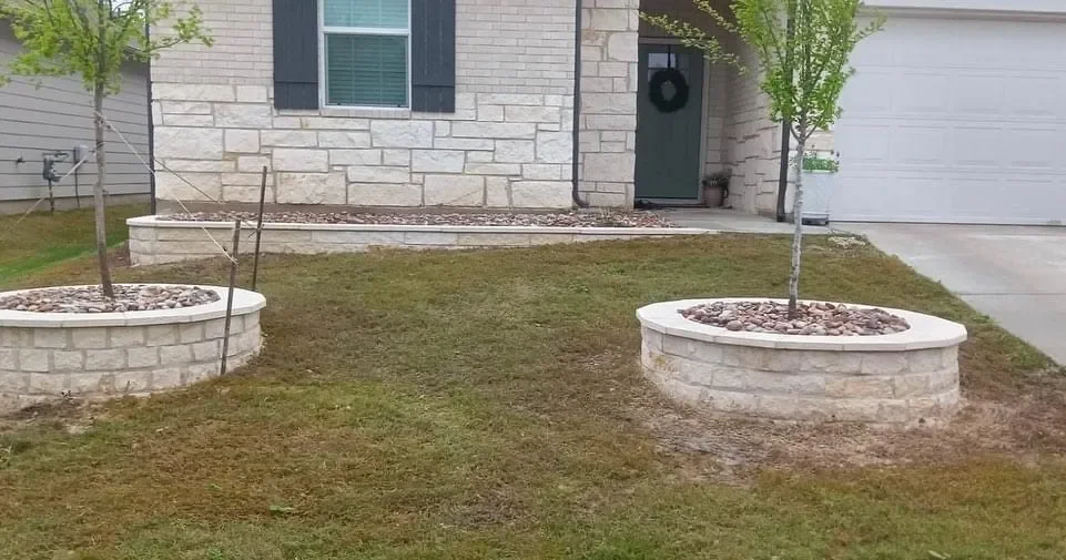 Front yard with two young trees in stone circular planters filled with rocks, a stone façade house with black shutters, a green door with a wreath, and a concrete driveway.